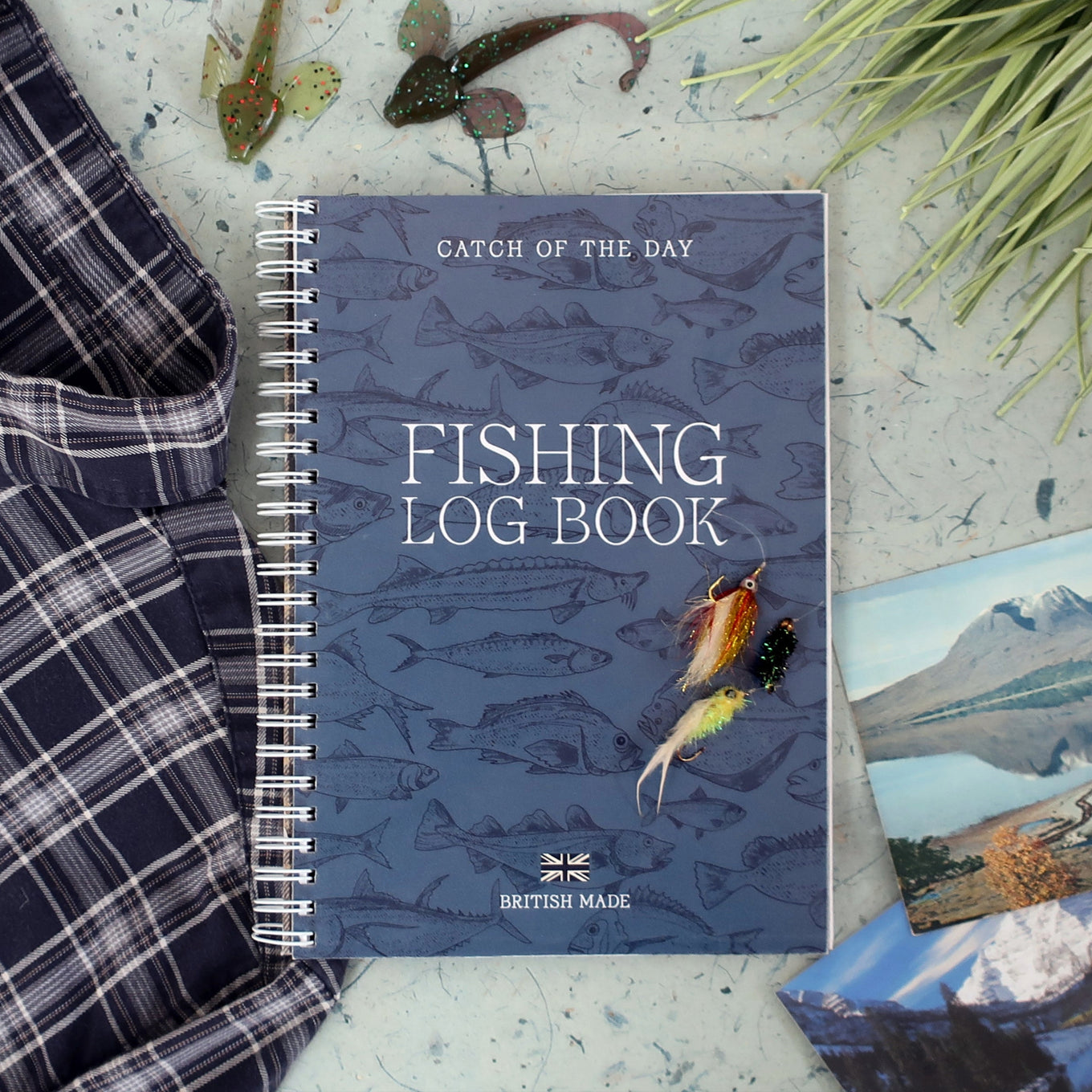 Fishing log book with fishing-themed cover on a surface with fishing equipment and a vintage scenic postcard
