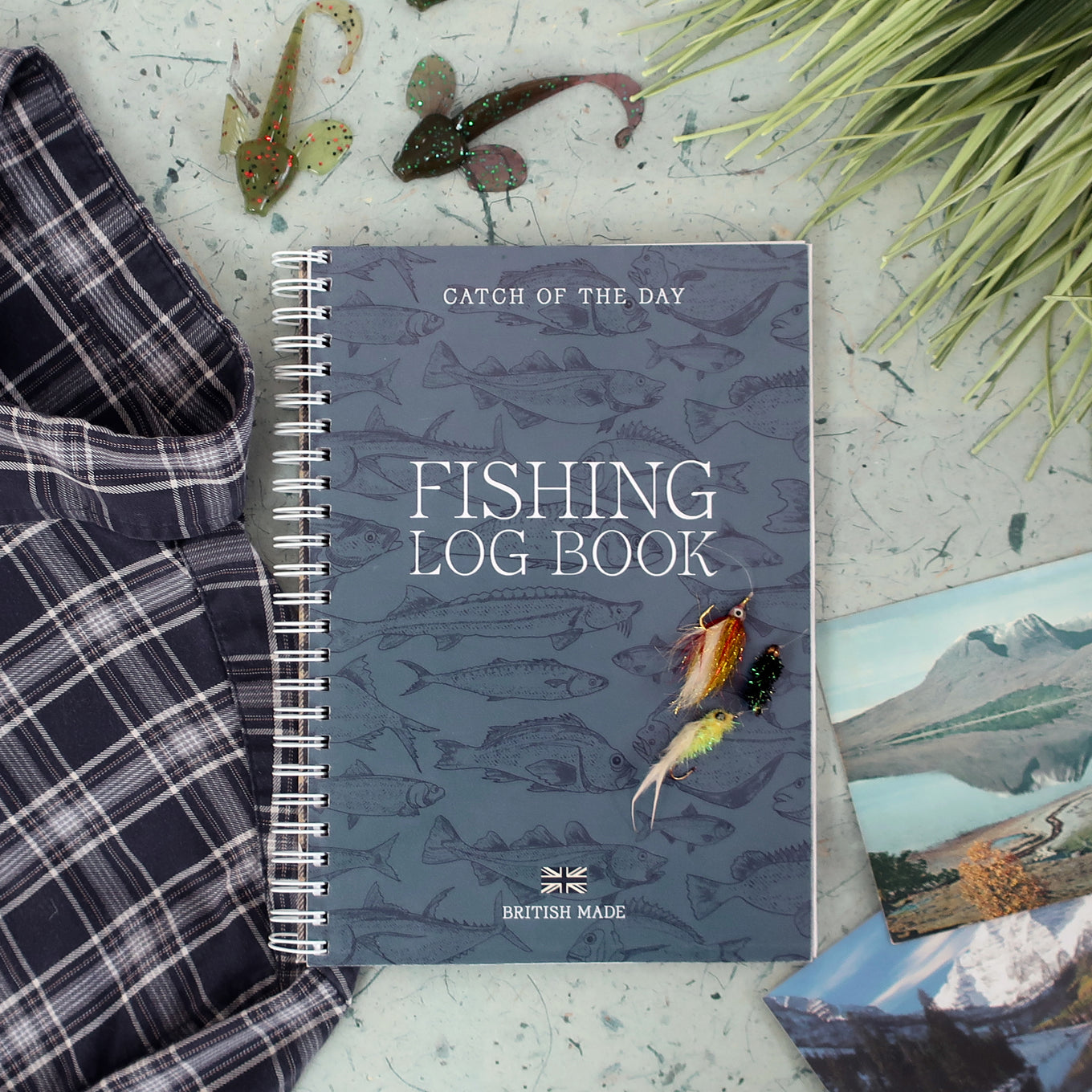 Fishing log book with fishing-themed cover on a surface with fishing equipment and a vintage scenic postcard
