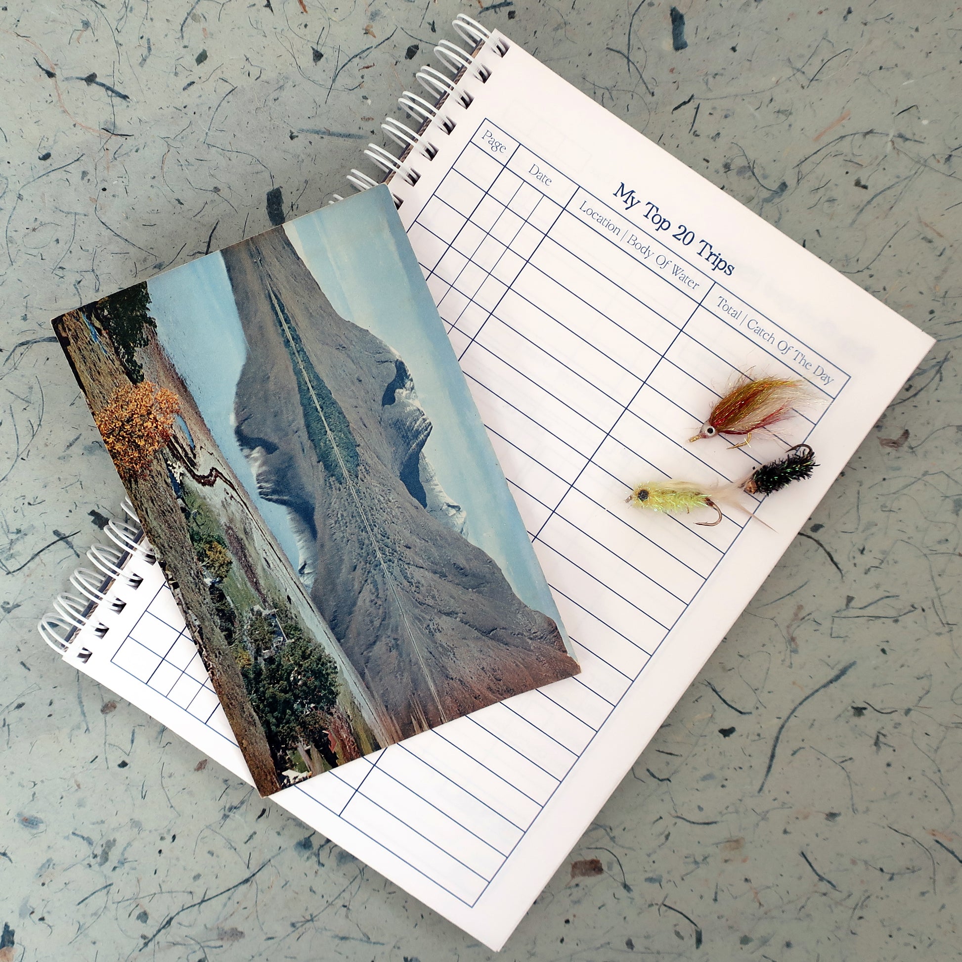 Fishing log book with my Top 20 Trips page open, with a mountain postcard, fishing flies laid on top on a textured surface
