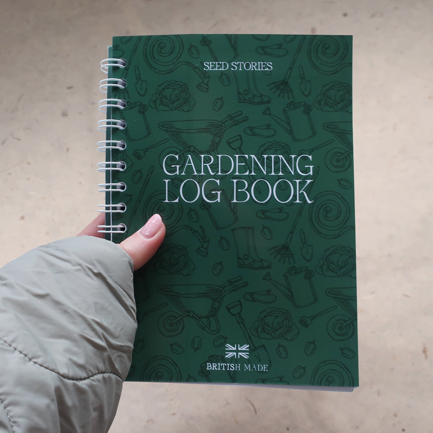 Gardening log book held by a hand wearing a green quilted gardening jacket on a beige background