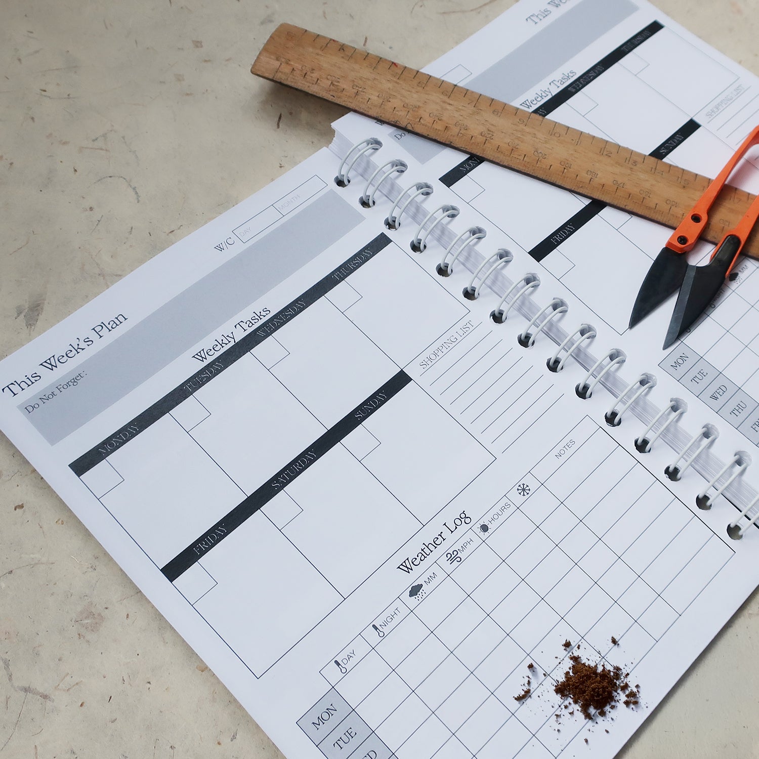Gardening Log Book open on the Weekly Planner page. With areas to log to the weather and weekly tasks. With a wooden ruler and garden shears laid over the plan