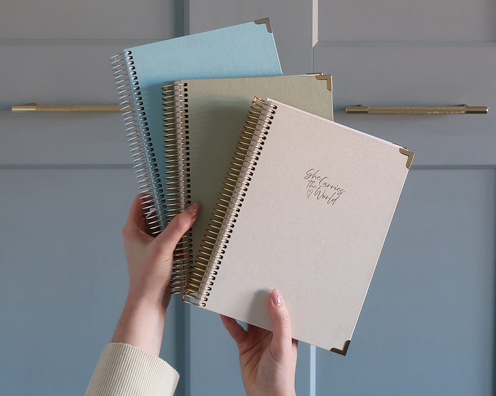 Hand holding three spiral-bound pregnancy journals with different coloured linen covers