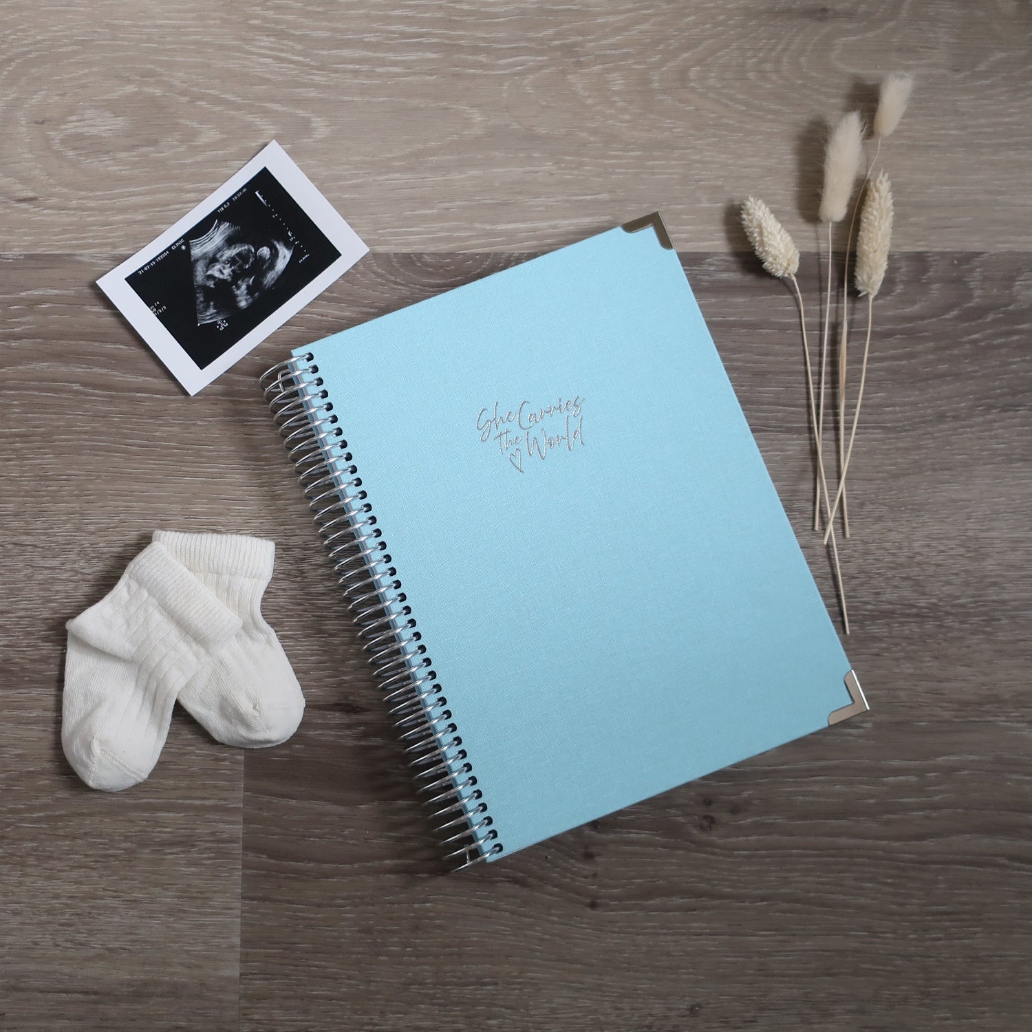 Light Blue pregnancy journal with silver spiral binding and 'She Carries the World' text on a light wooden desk, with ultrasound photograph and baby cream socks