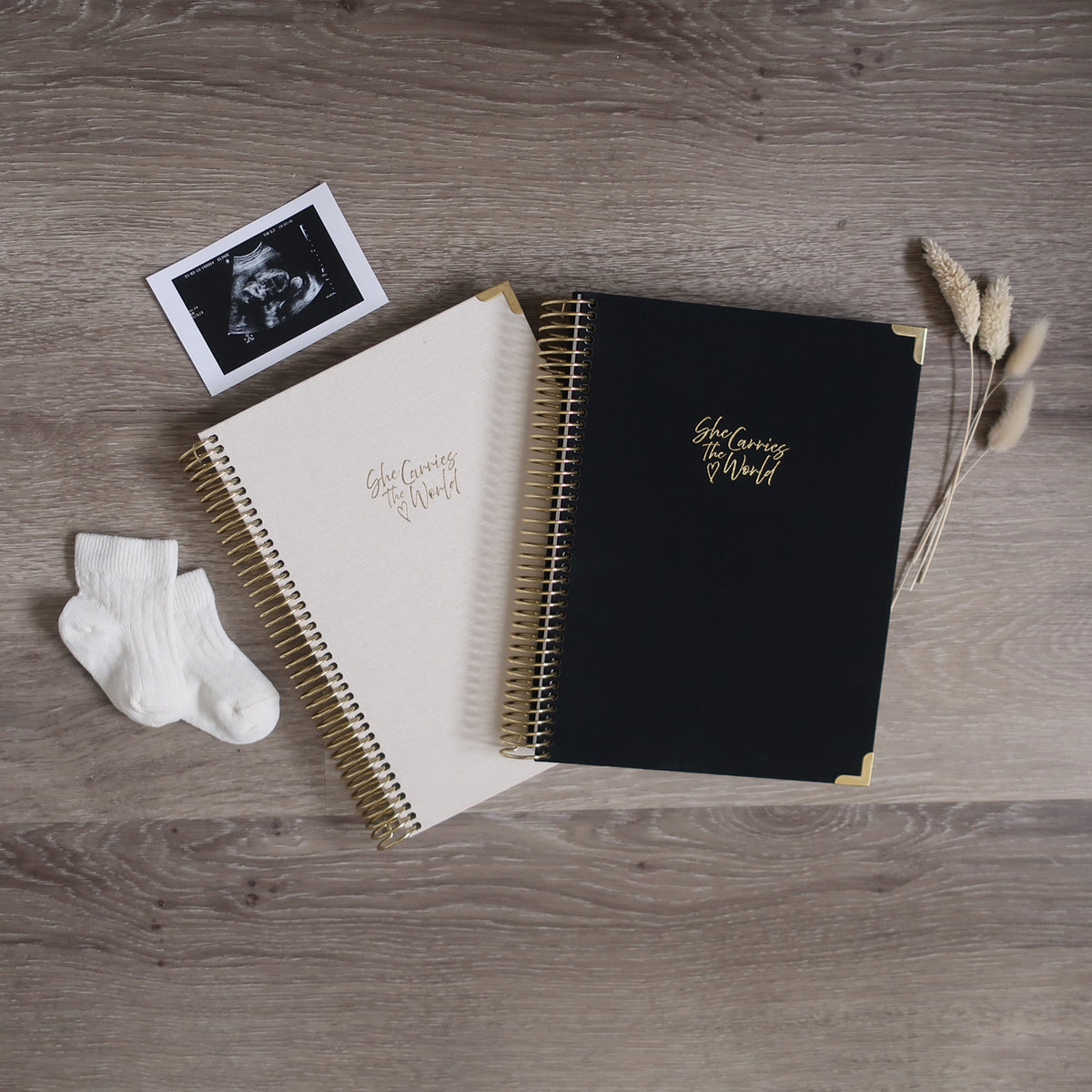 Two Pregnancy Journals On A Wooden Background Surrounded By A Ultrasound & Baby Socks. Ring Bound Pregnancy Journals With Linen Covers One Cream & One Black