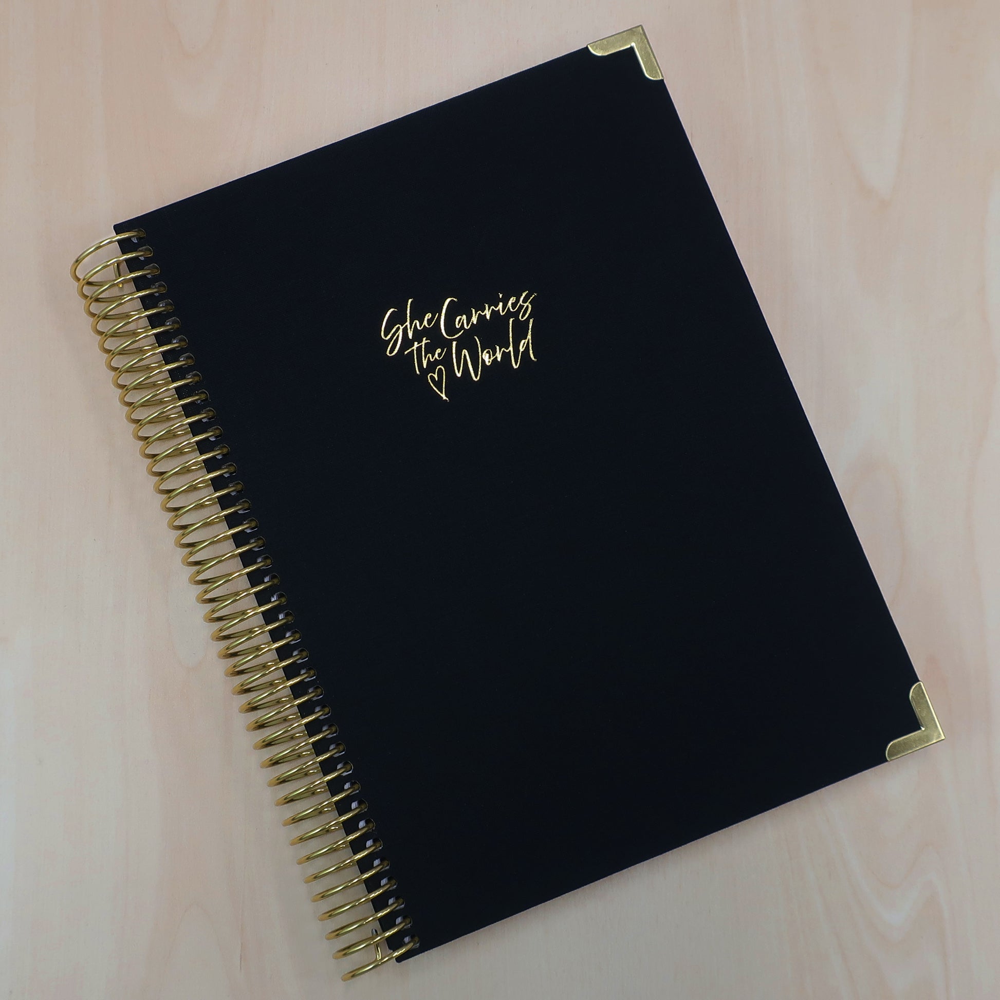 Black pregnancy journal with gold spiral binding and 'She Carries the World' text on a light wooden desk