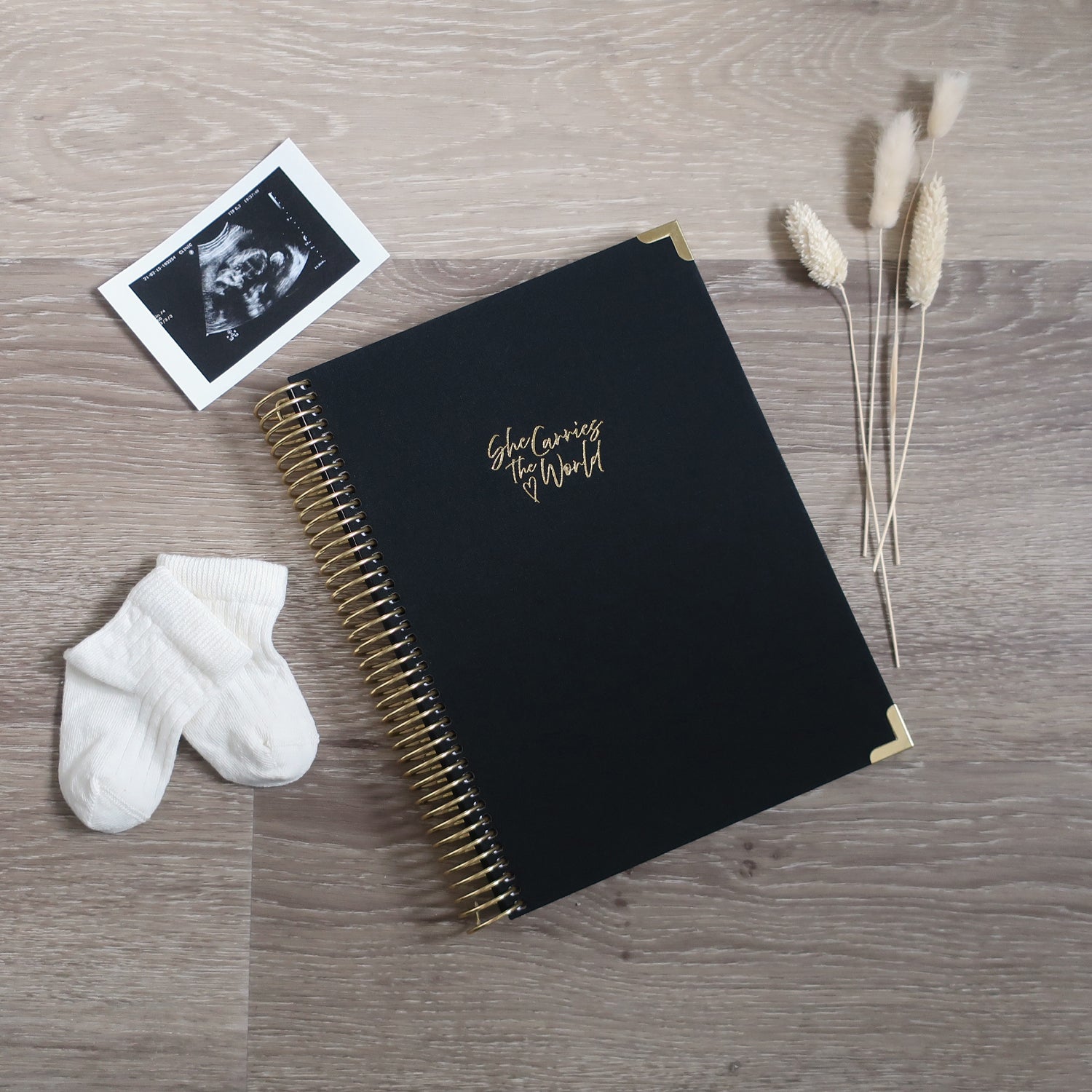 Black pregnancy journal with gold spiral binding and 'She Carries the World' text on a light wooden desk, with ultrasound photograph and baby cream socks