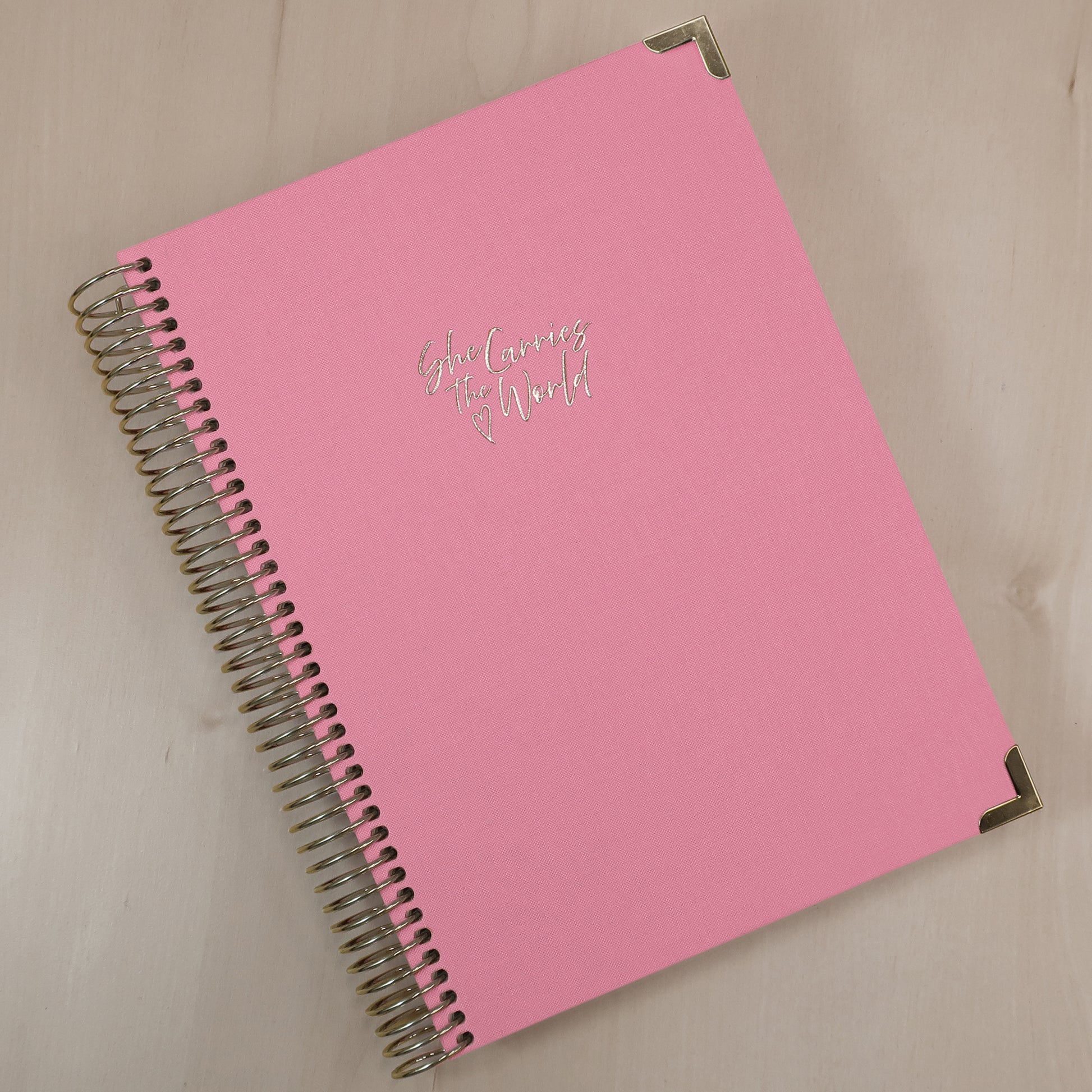 Baby Pink pregnancy journal with gold spiral binding and 'She Carries the World' text on a light wooden desk