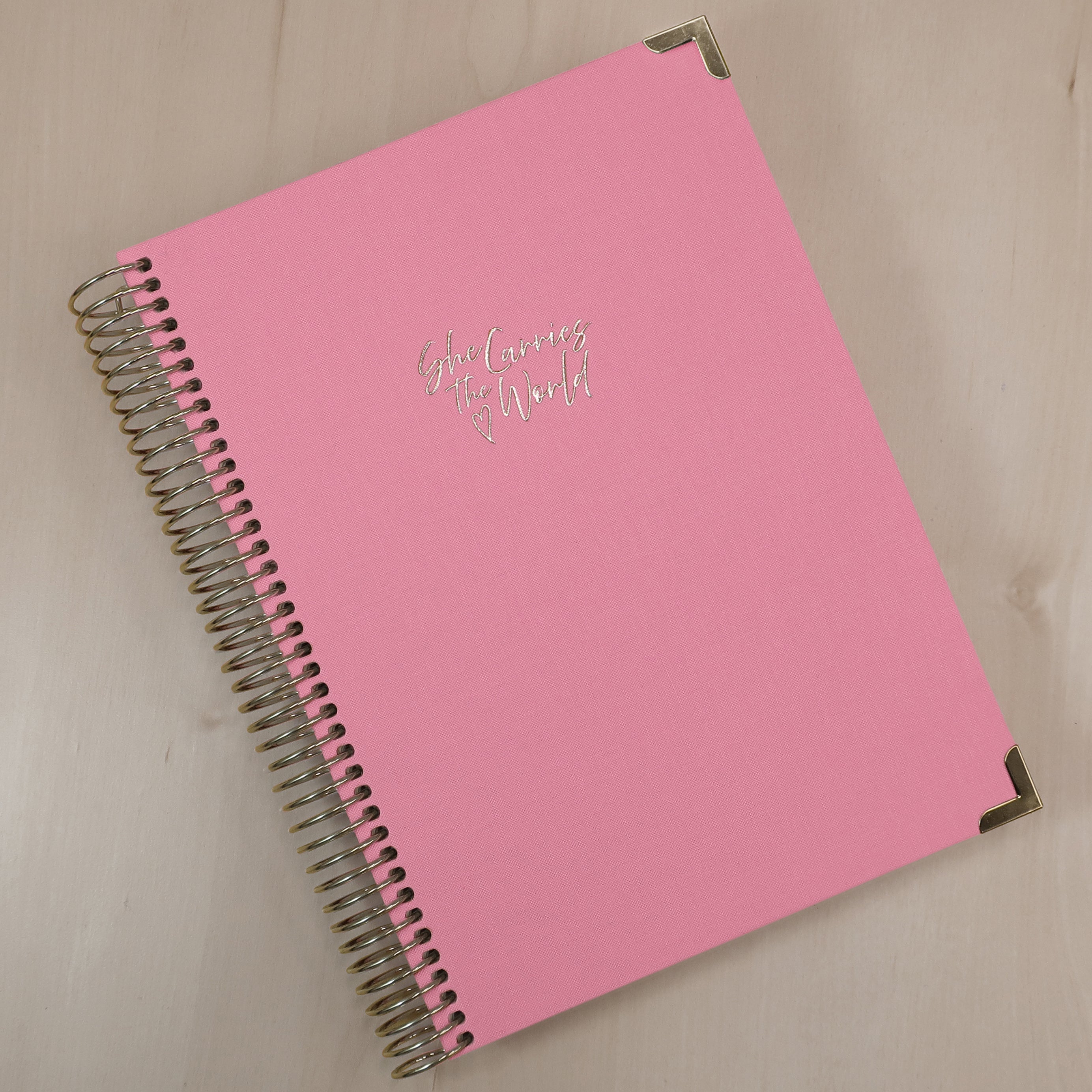 Baby Pink pregnancy journal with gold spiral binding and 'She Carries the World' text on a light wooden desk