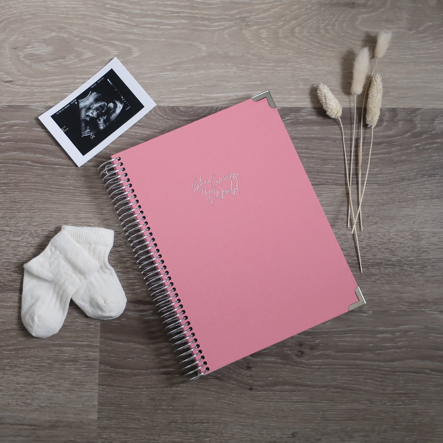 Baby Pink pregnancy journal with Silver spiral binding and 'She Carries the World' text on a light wooden desk, with ultrasound photograph and baby cream socks