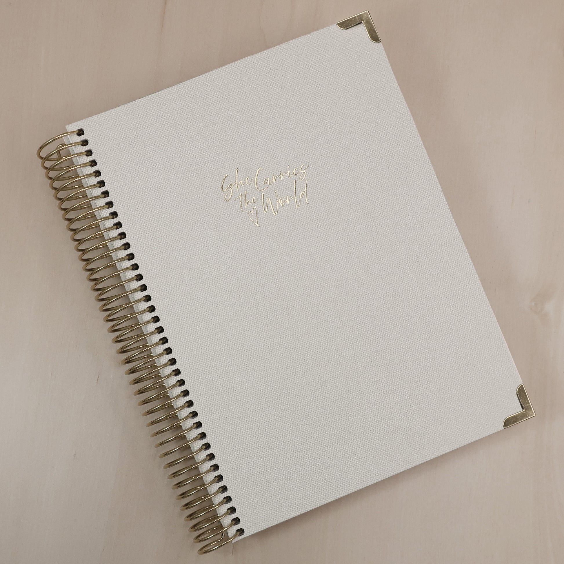 Cream pregnancy journal with gold spiral binding and 'She Carries the World' text on a light wooden desk