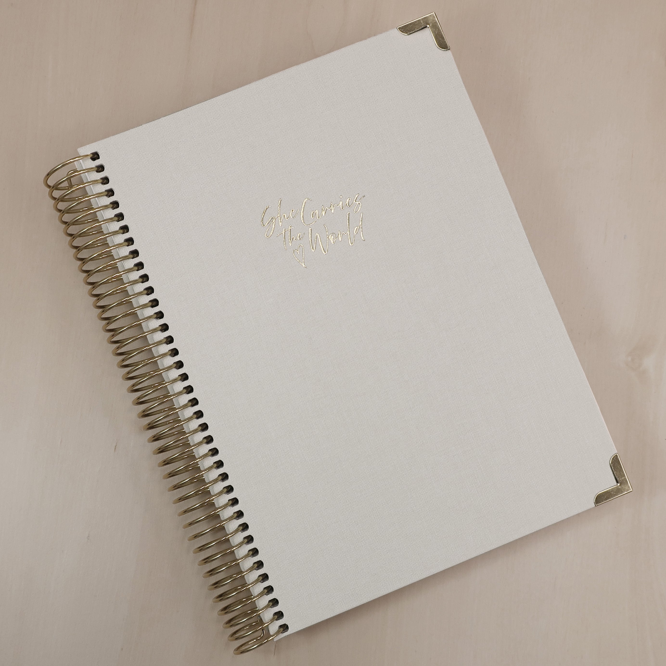 Cream pregnancy journal with gold spiral binding and 'She Carries the World' text on a light wooden desk