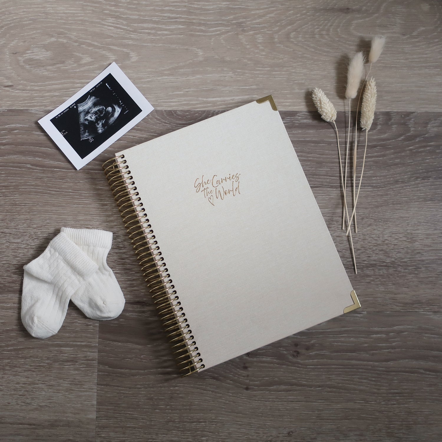 Cream pregnancy journal with gold spiral binding and 'She Carries the World' text on a light wooden desk, with ultrasound photograph and baby cream socks