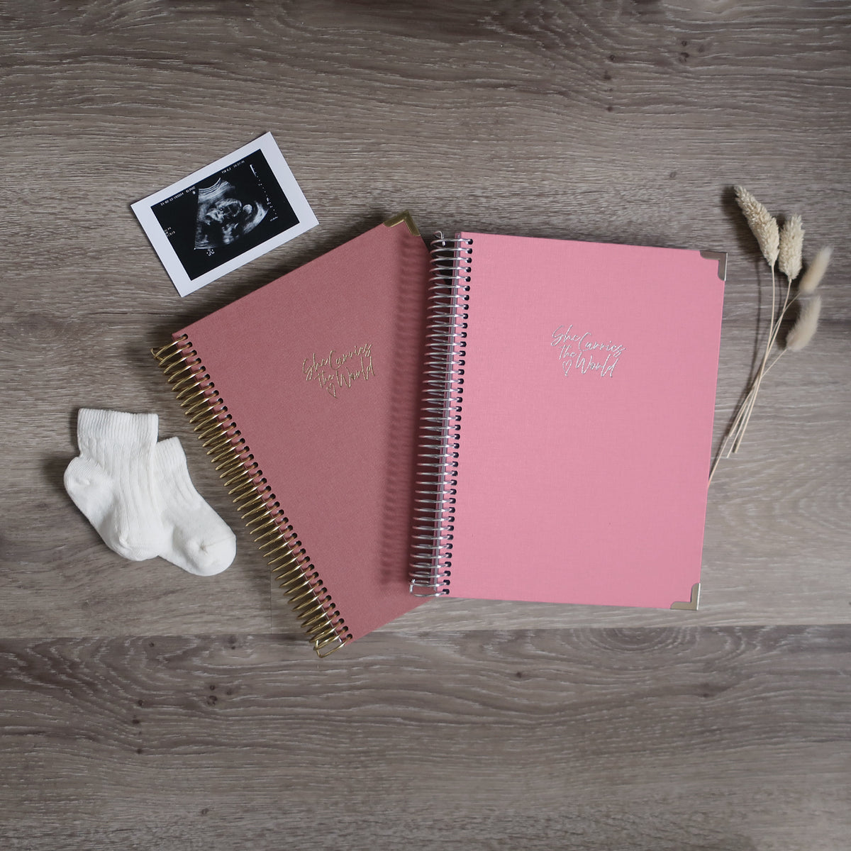 Two Pregnancy Journals On A Wooden Background Surrounded By A Ultrasound & Baby Socks. Ring Bound Pregnancy Journals With Terracotta & Baby Pink Linen Covers.