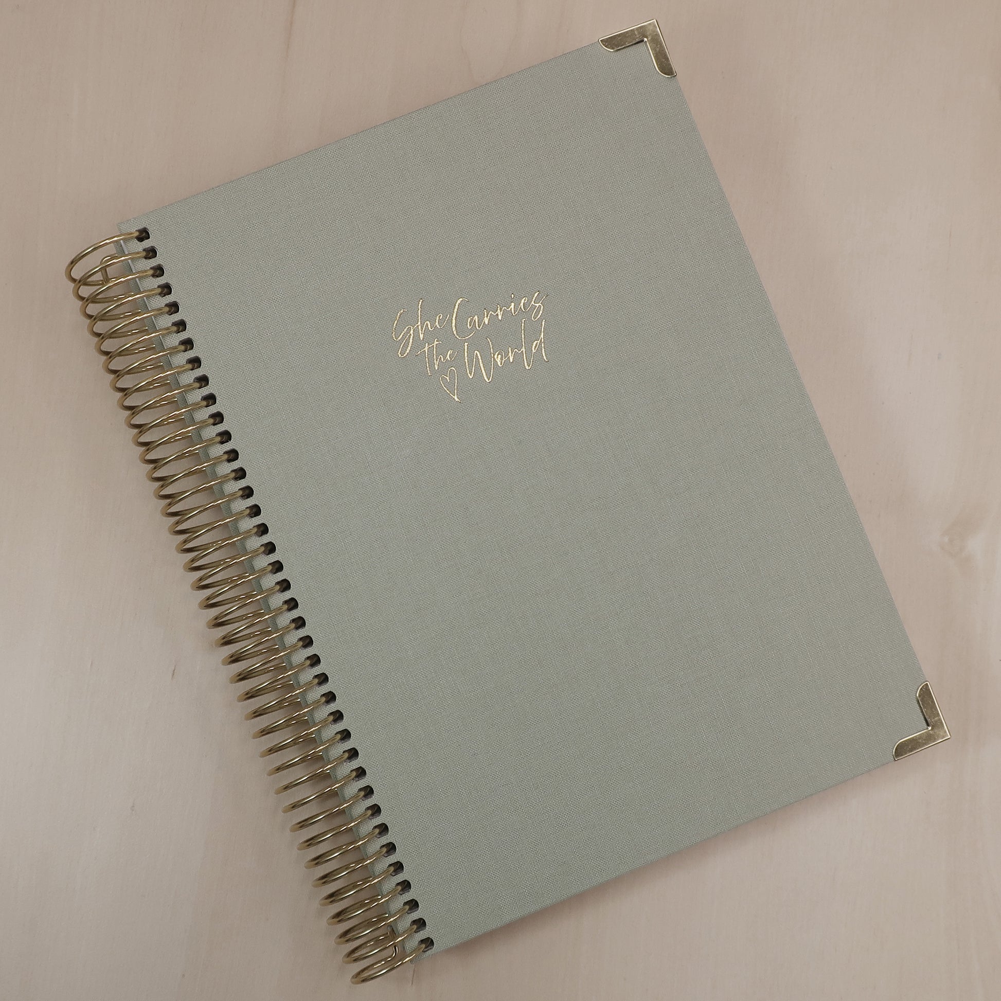 Pistachio Green pregnancy journal with gold spiral binding and 'She Carries the World' text on a light wooden desk