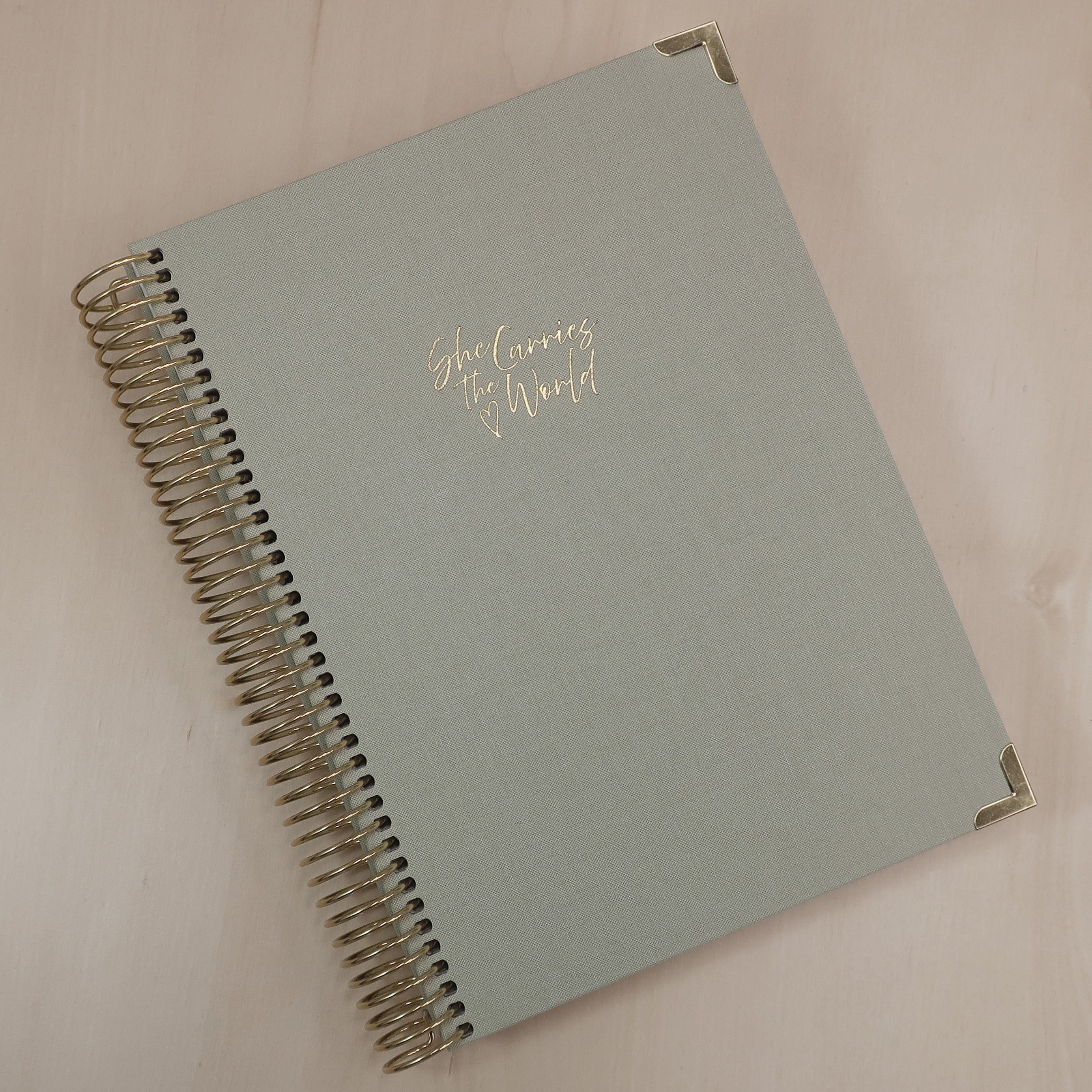 Pistachio Green pregnancy journal with gold spiral binding and 'She Carries the World' text on a light wooden desk