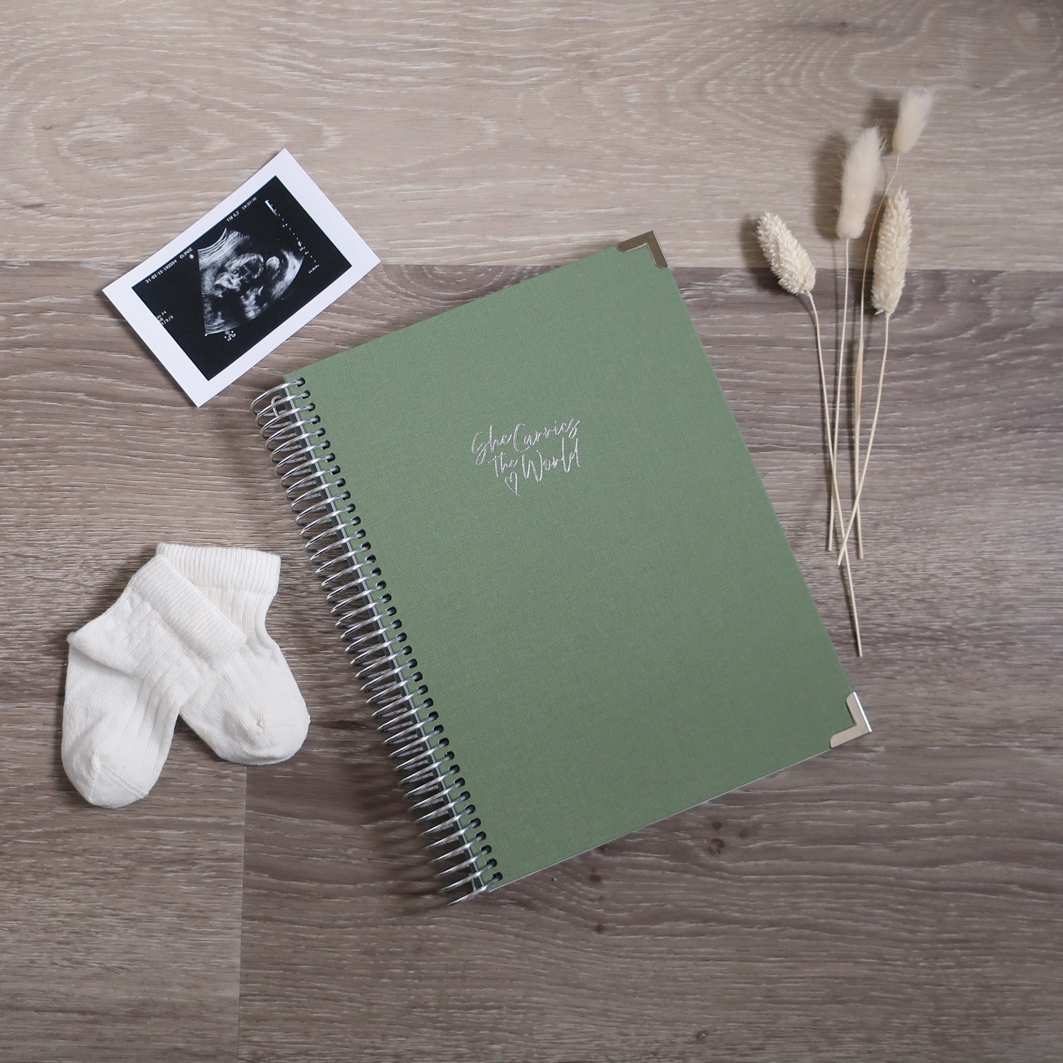 Sage Green pregnancy journal with Silver spiral binding and 'She Carries the World' text on a light wooden desk, with ultrasound photograph and baby cream socks