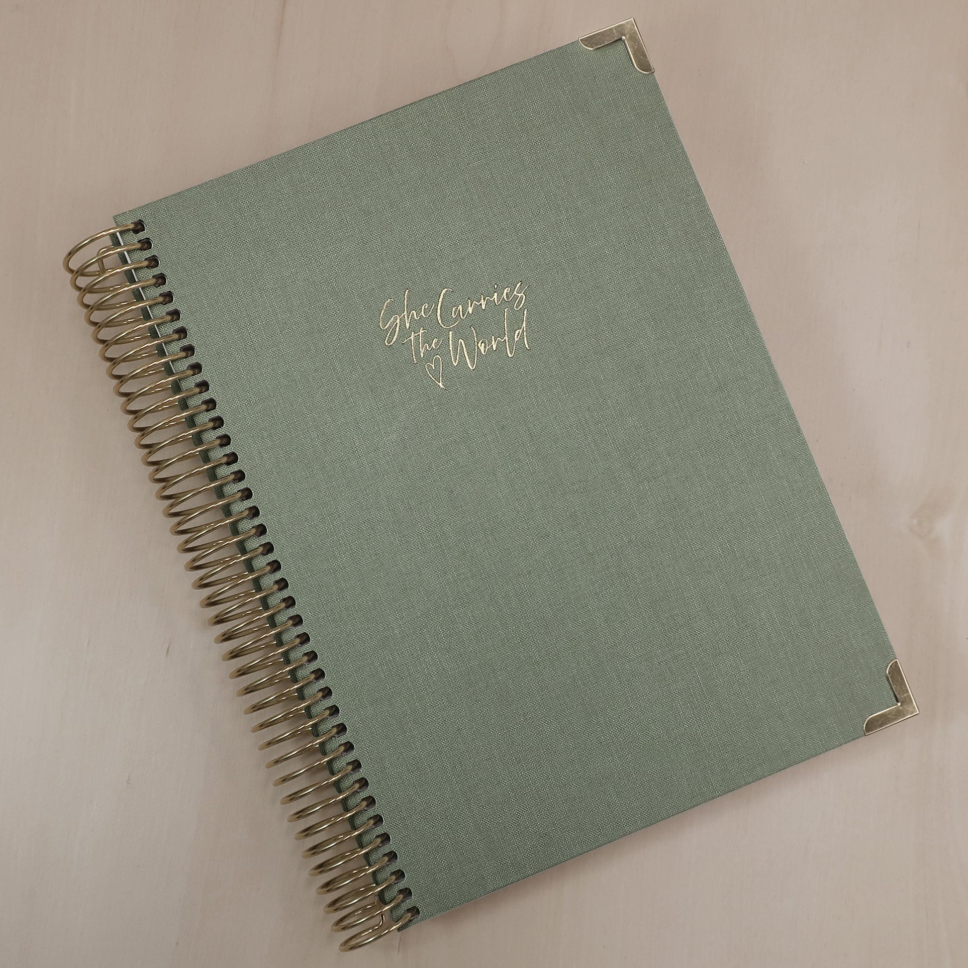 Sage Green pregnancy journal with gold spiral binding and 'She Carries the World' text on a light wooden desk