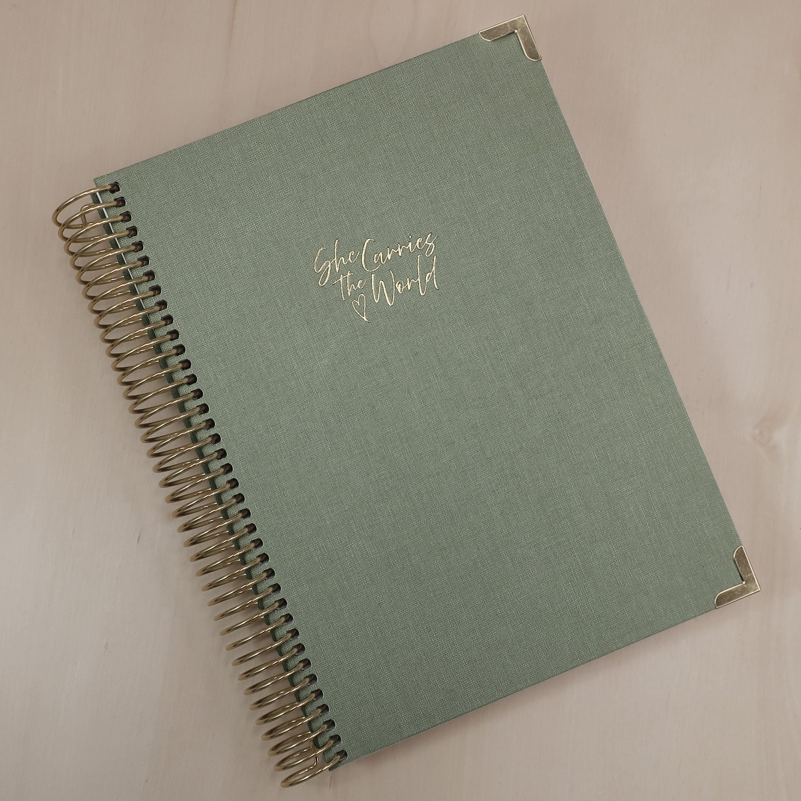 Sage Green pregnancy journal with gold spiral binding and 'She Carries the World' text on a light wooden desk