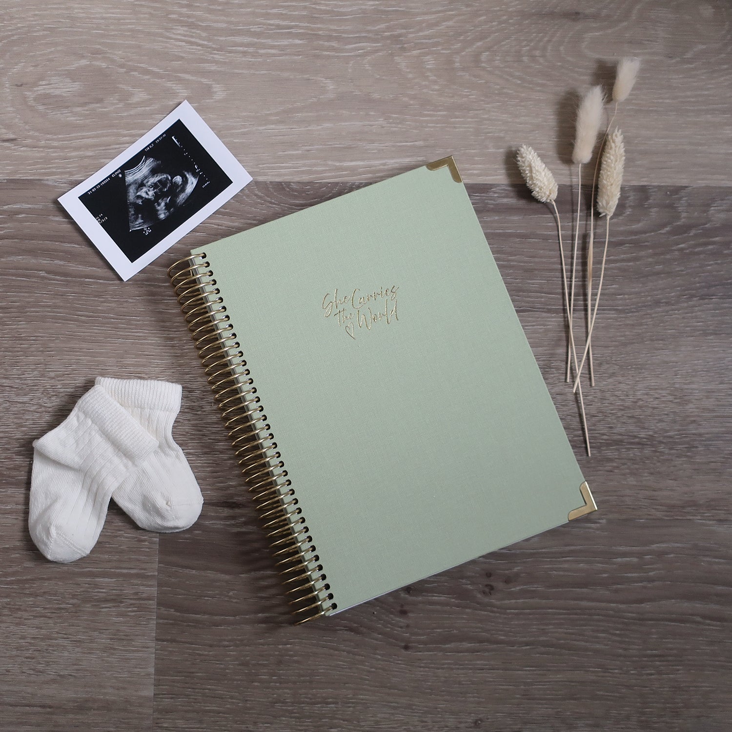 Pistachio Green pregnancy journal with gold spiral binding and 'She Carries the World' text on a light wooden desk, with ultrasound photograph and baby cream socks