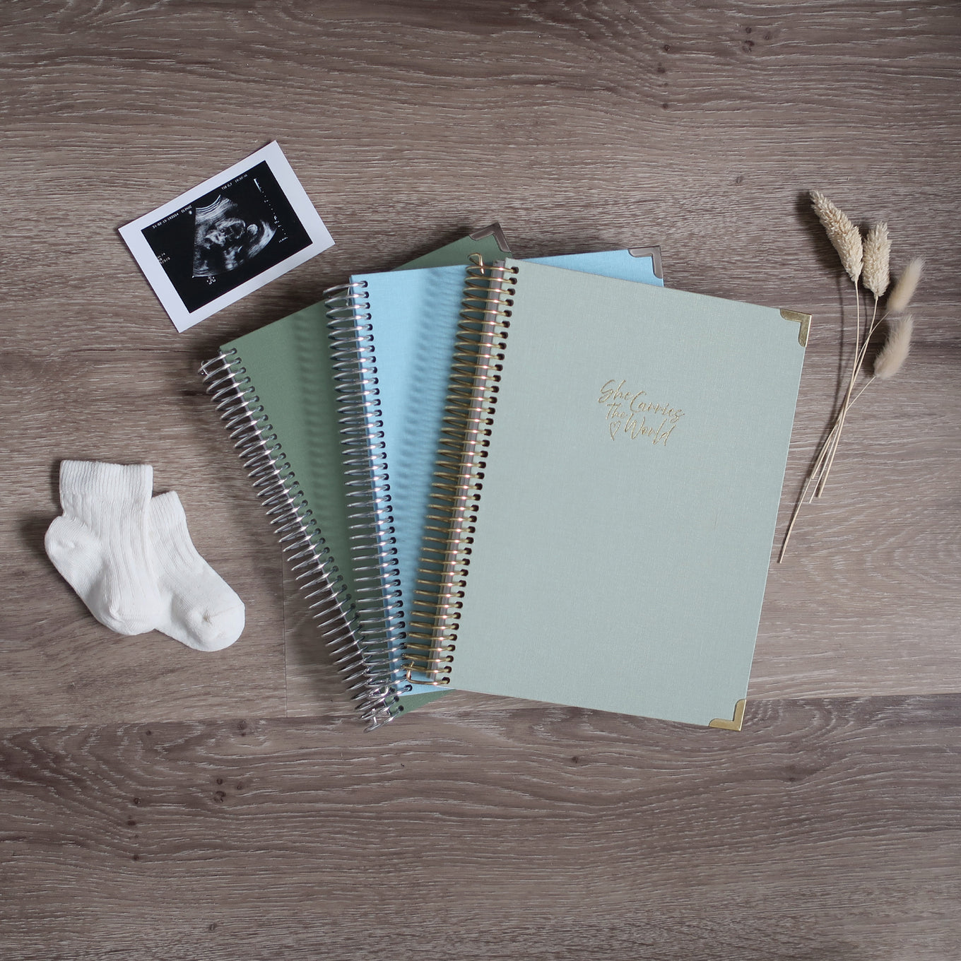 Three Pregnancy Journals On A Wooden Background Surrounded By A Ultrasound & Baby Socks. Ring Bound Pregnancy Journals With Linen Covers One Sage Green, One Baby Blue & One Pistachio