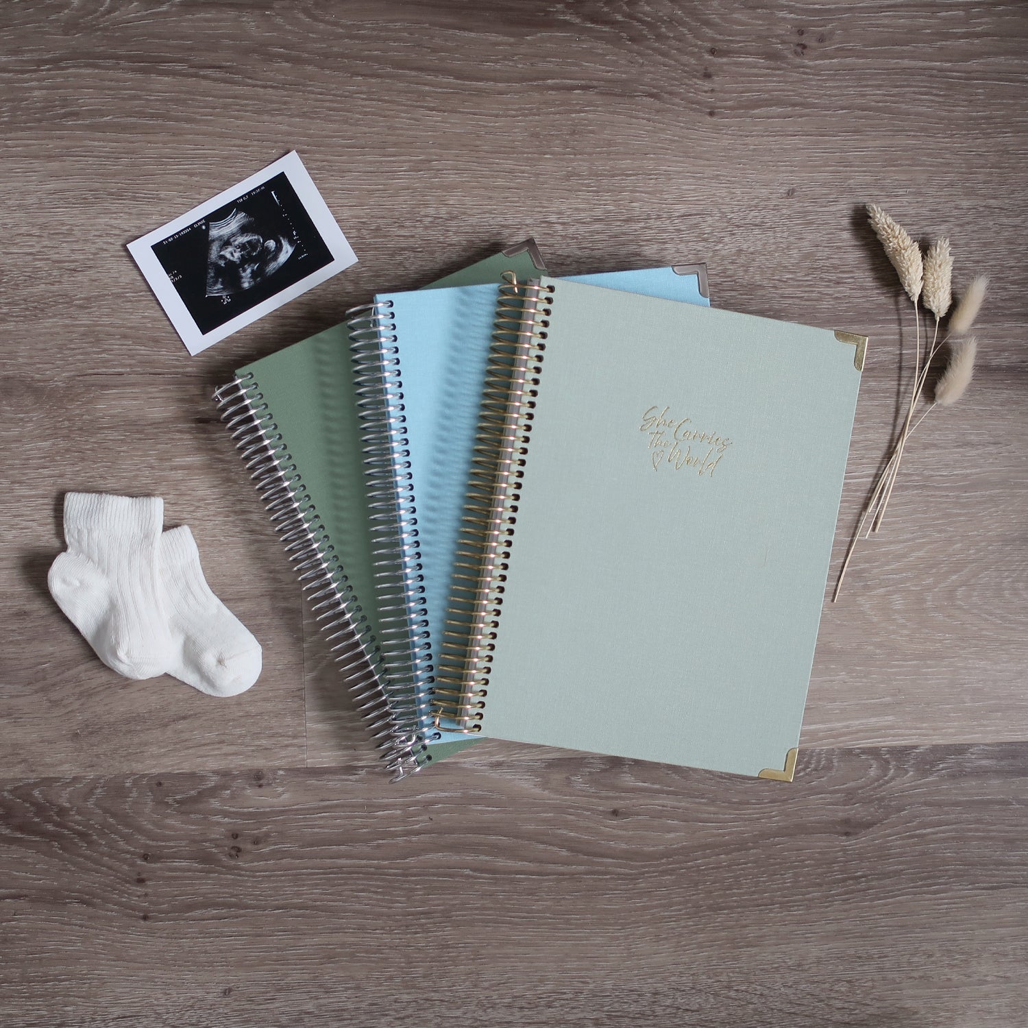 Three Pregnancy Journals On A Wooden Background Surrounded By A Ultrasound & Baby Socks. Ring Bound Pregnancy Journals With Linen Covers One Sage Green, One Baby Blue & One Pistachio