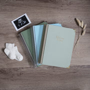 Three Pregnancy Journals On A Wooden Background Surrounded By A Ultrasound & Baby Socks. Ring Bound Pregnancy Journals With Linen Covers One Sage Green, One Baby Blue & One Pistachio