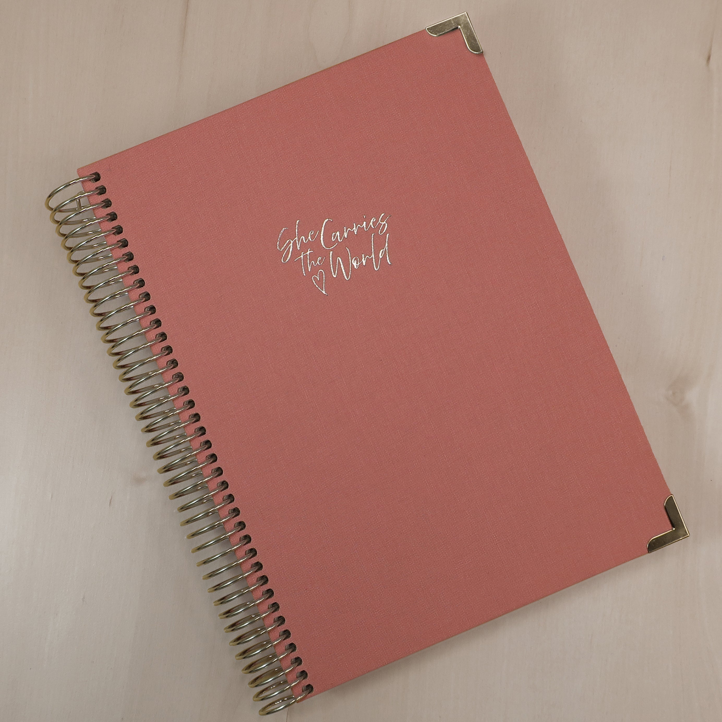 Terracotta Rose pregnancy journal with gold spiral binding and 'She Carries the World' text on a light wooden desk