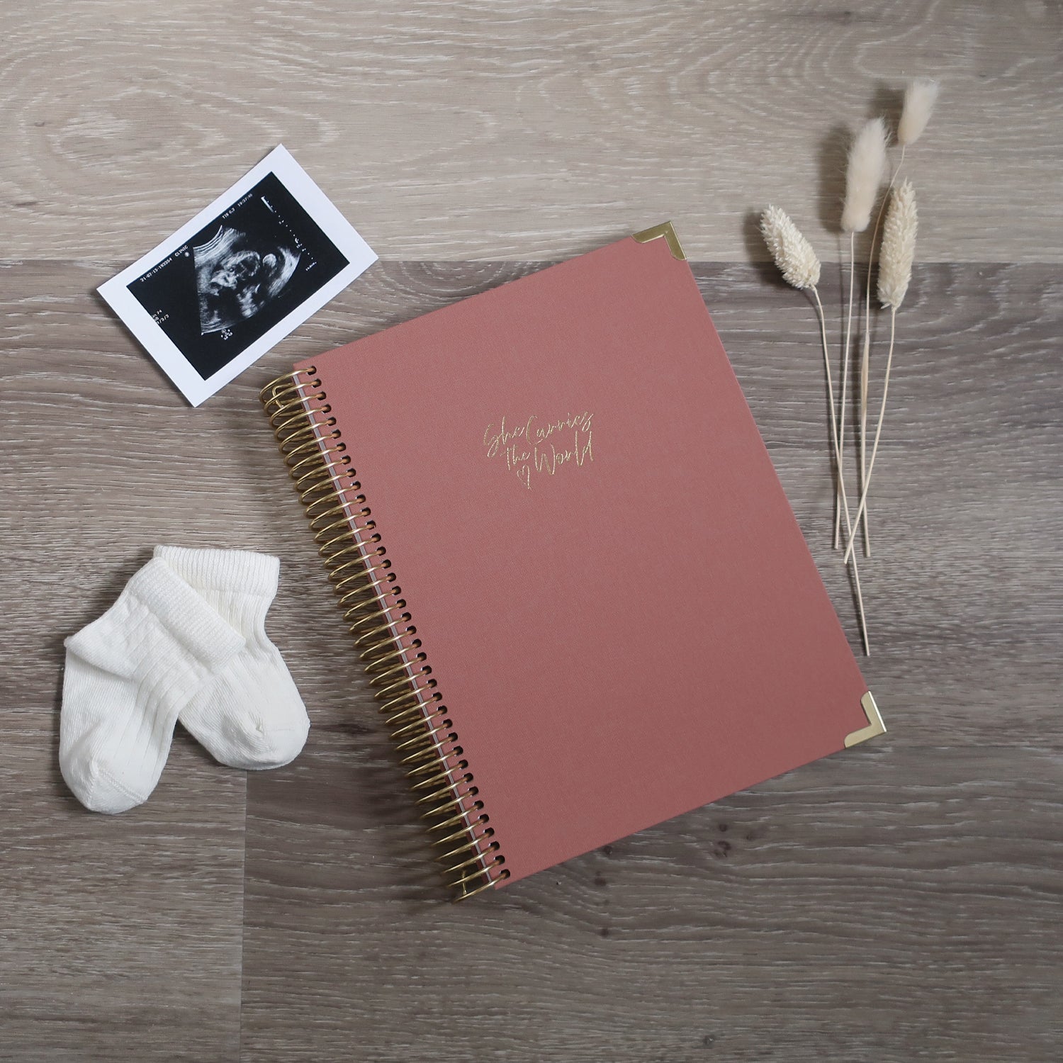 Terracotta Rose pregnancy journal with gold spiral binding and 'She Carries the World' text on a light wooden desk, with ultrasound photograph and baby cream socks