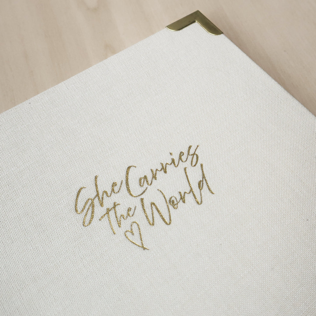 Cream linen pregnancy journal with gold text 'She Carries The World' on a wooden desk