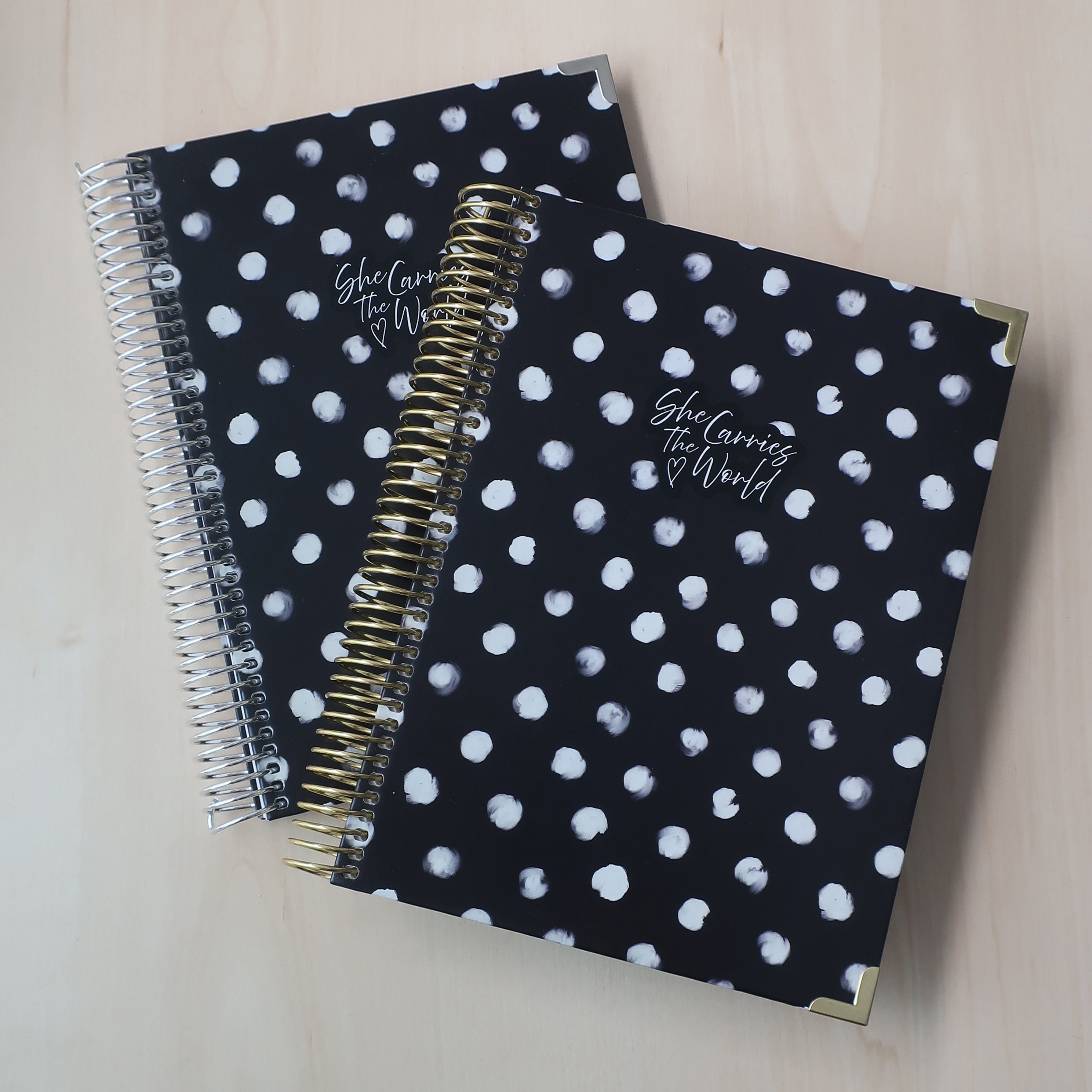 Two spiral-bound pregnancy journals with black and white polka dot pattern on a wooden desk. With the 'She Carries The World' logo printed to the cover. 