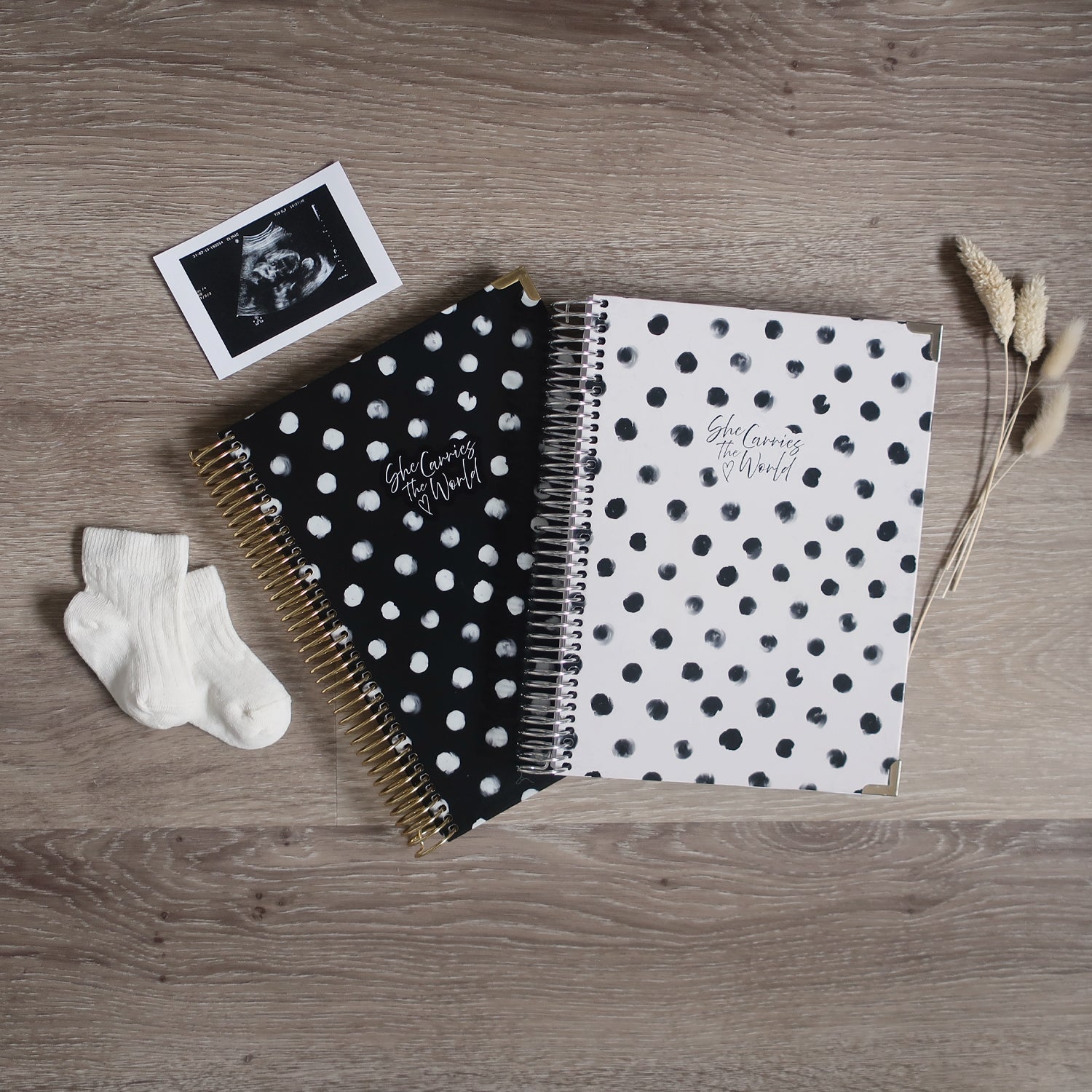 Two Pregnancy Journals On A Wooden Background Surrounded By A Ultrasound & Baby Socks. Ring Bound Pregnancy Journals With One Black Polka Dot Cover, One White Polka Dot Cover.
