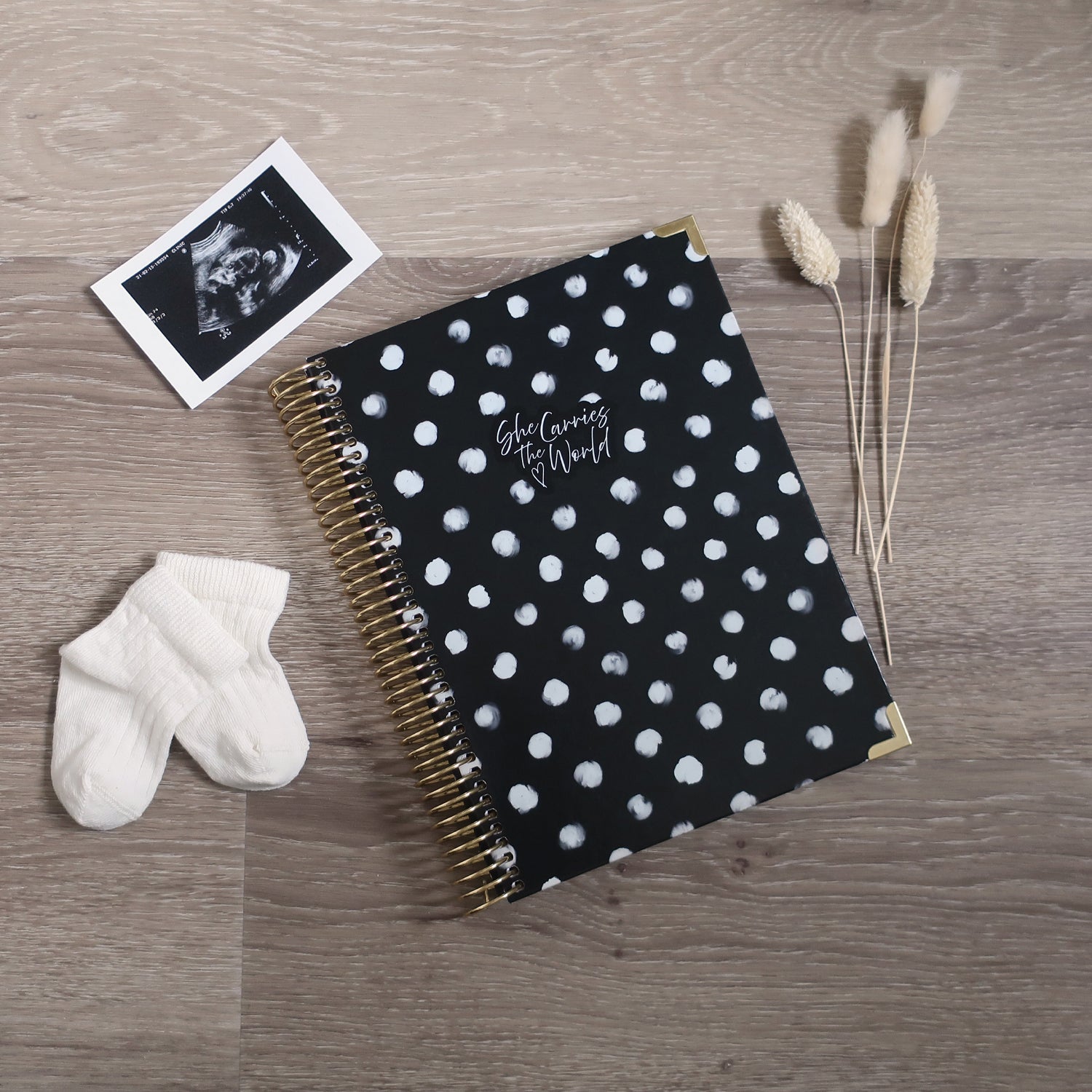 Black spiral-bound pregnancy journal with white polka dots on a wooden desk, next to a ultrasound photo and baby socks.