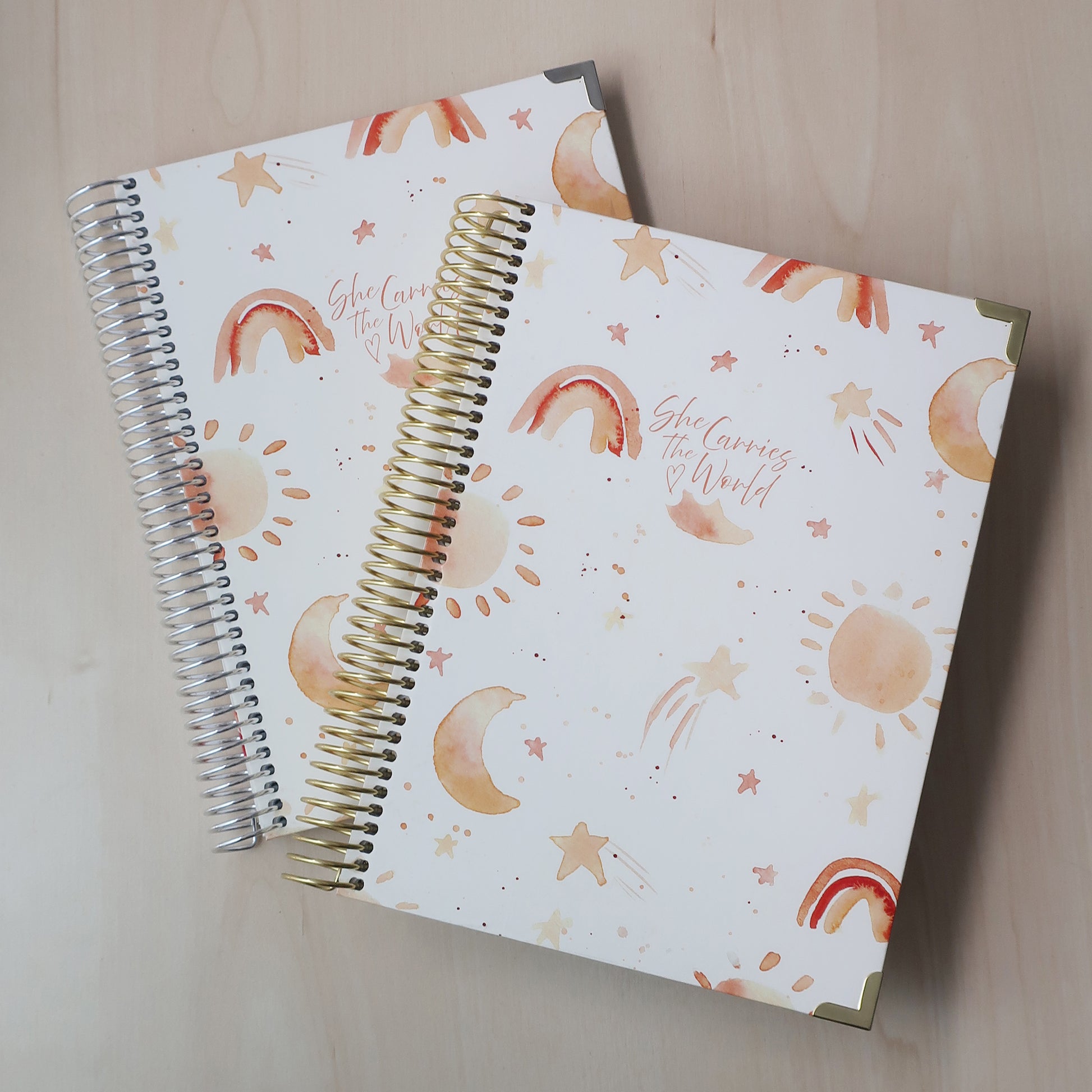 Two spiral-bound pregnancy journals with water coloured suns, stars and moons pattern on a wooden desk. With the 'She Carries The World' logo printed to the cover. 