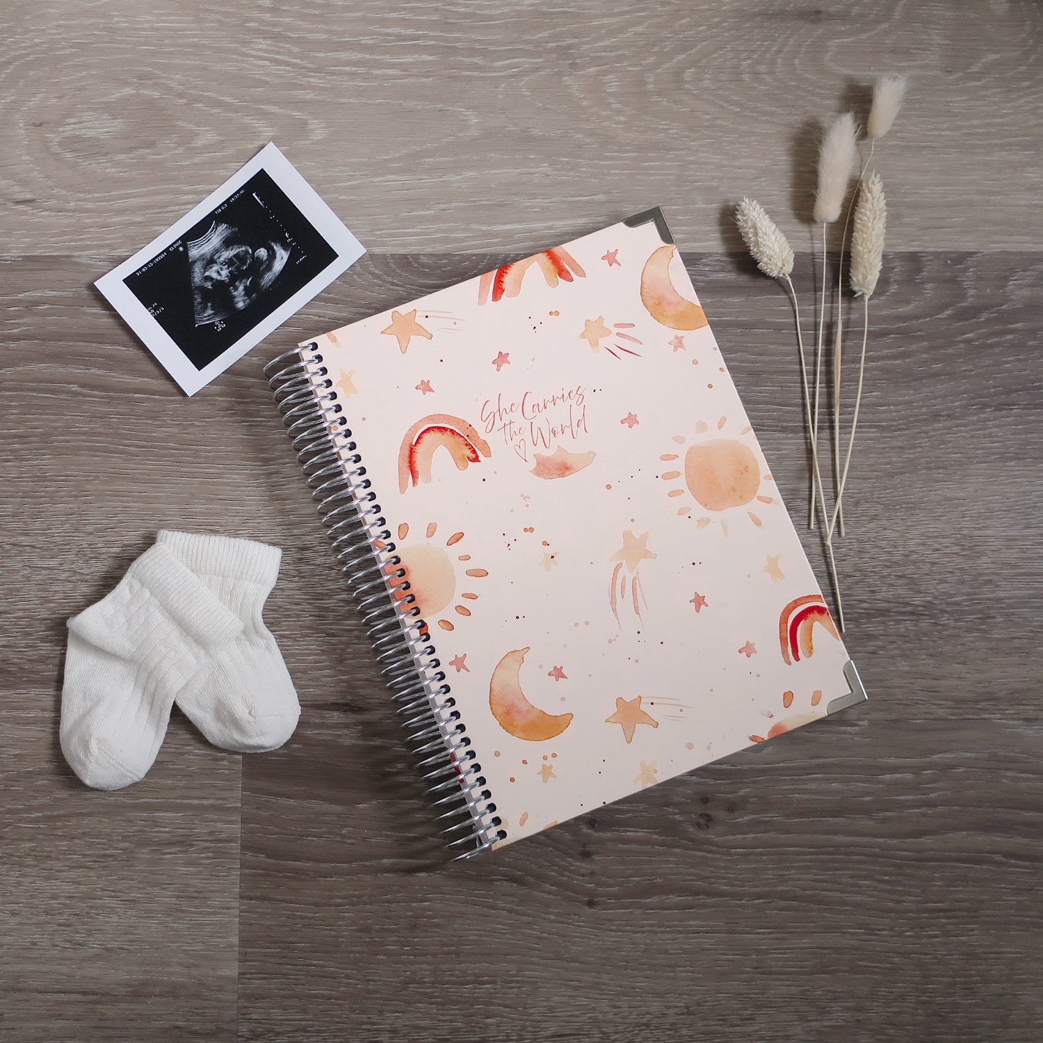 Baby themed watercoloured printed spiral-bound pregnancy journal on a wooden desk, next to a ultrasound photo and baby socks.