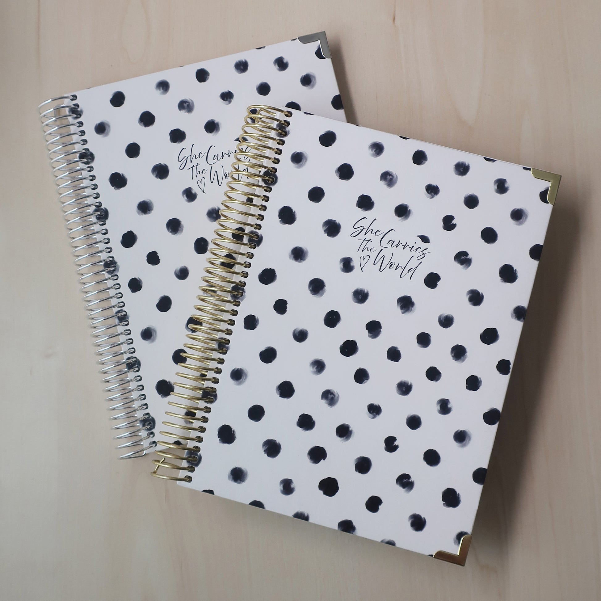 Two spiral-bound pregnancy journals with white and black polka dot pattern on a wooden desk. With the 'She Carries The World' logo printed to the cover. 