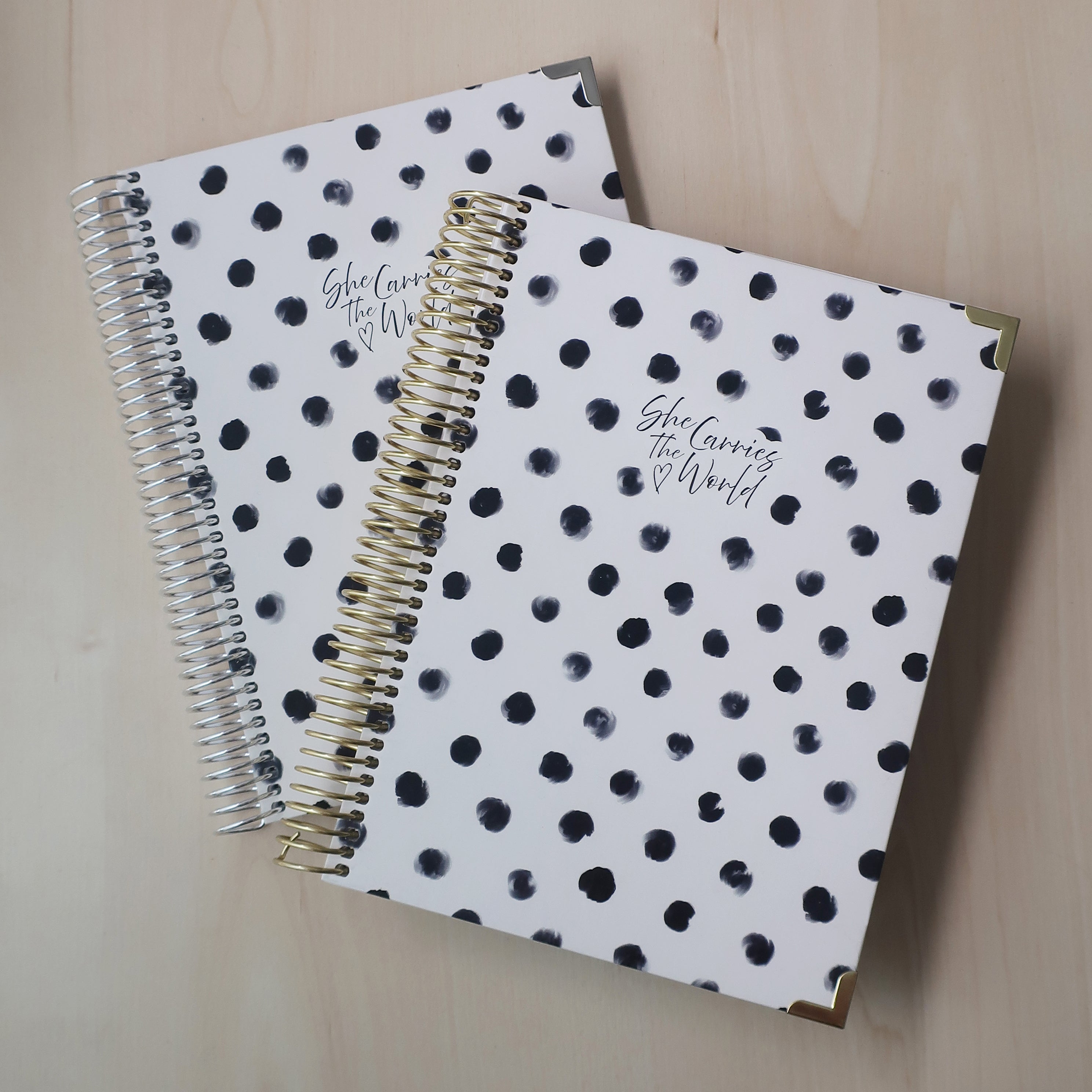 Two spiral-bound pregnancy journals with white and black polka dot pattern on a wooden desk. With the 'She Carries The World' logo printed to the cover. 