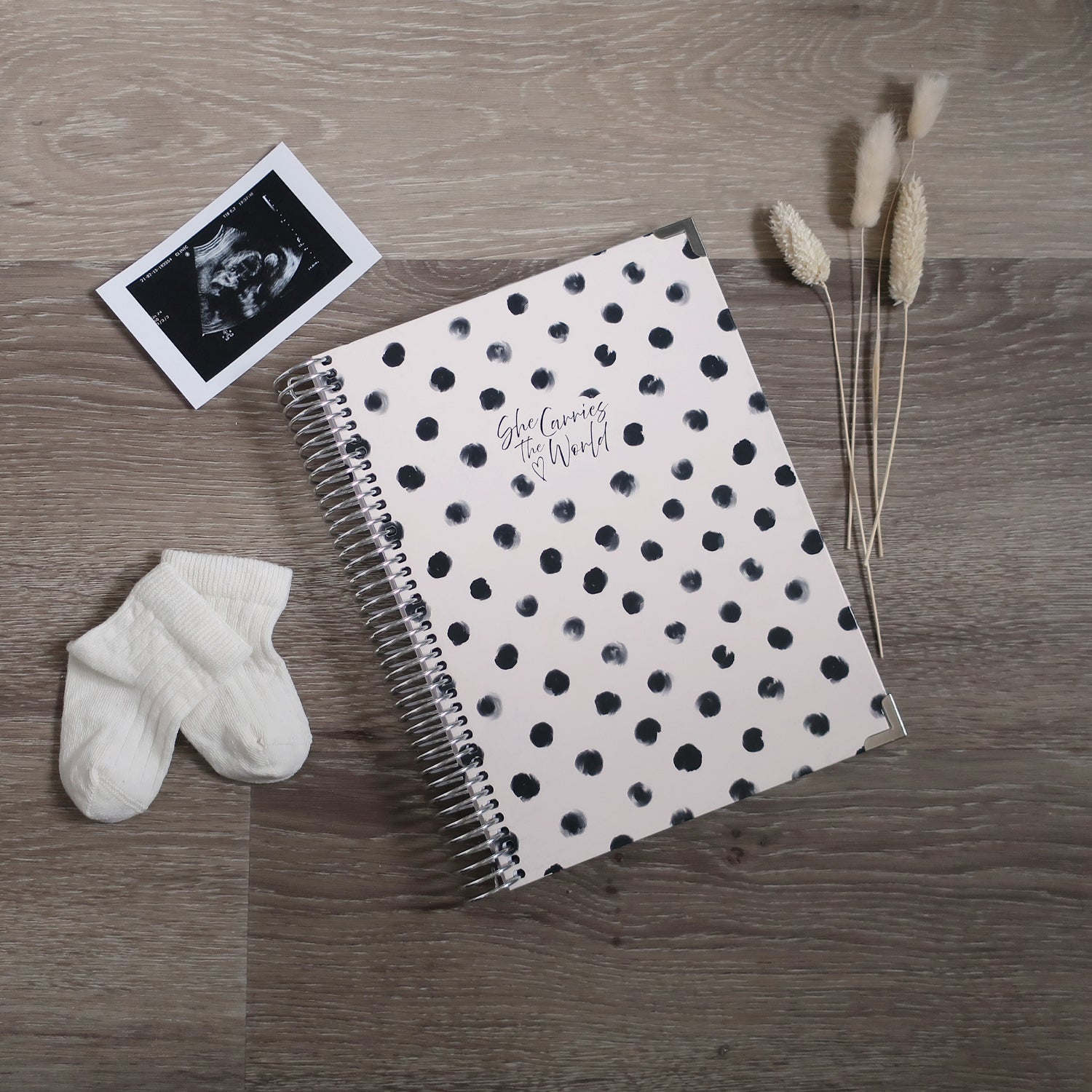 White spiral-bound pregnancy journal with white polka dots on a wooden desk, next to a ultrasound photo and baby socks.