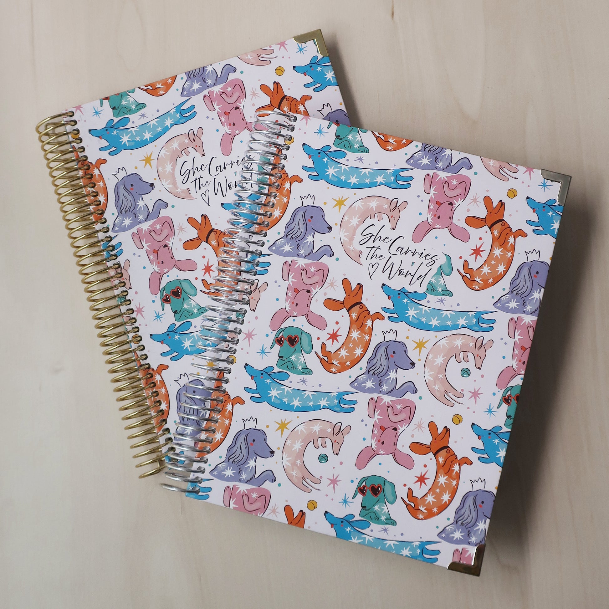 Two spiral-bound pregnancy journals with abstract colourful dachshund pattern on a wooden desk. With the 'She Carries The World' logo printed to the cover. 