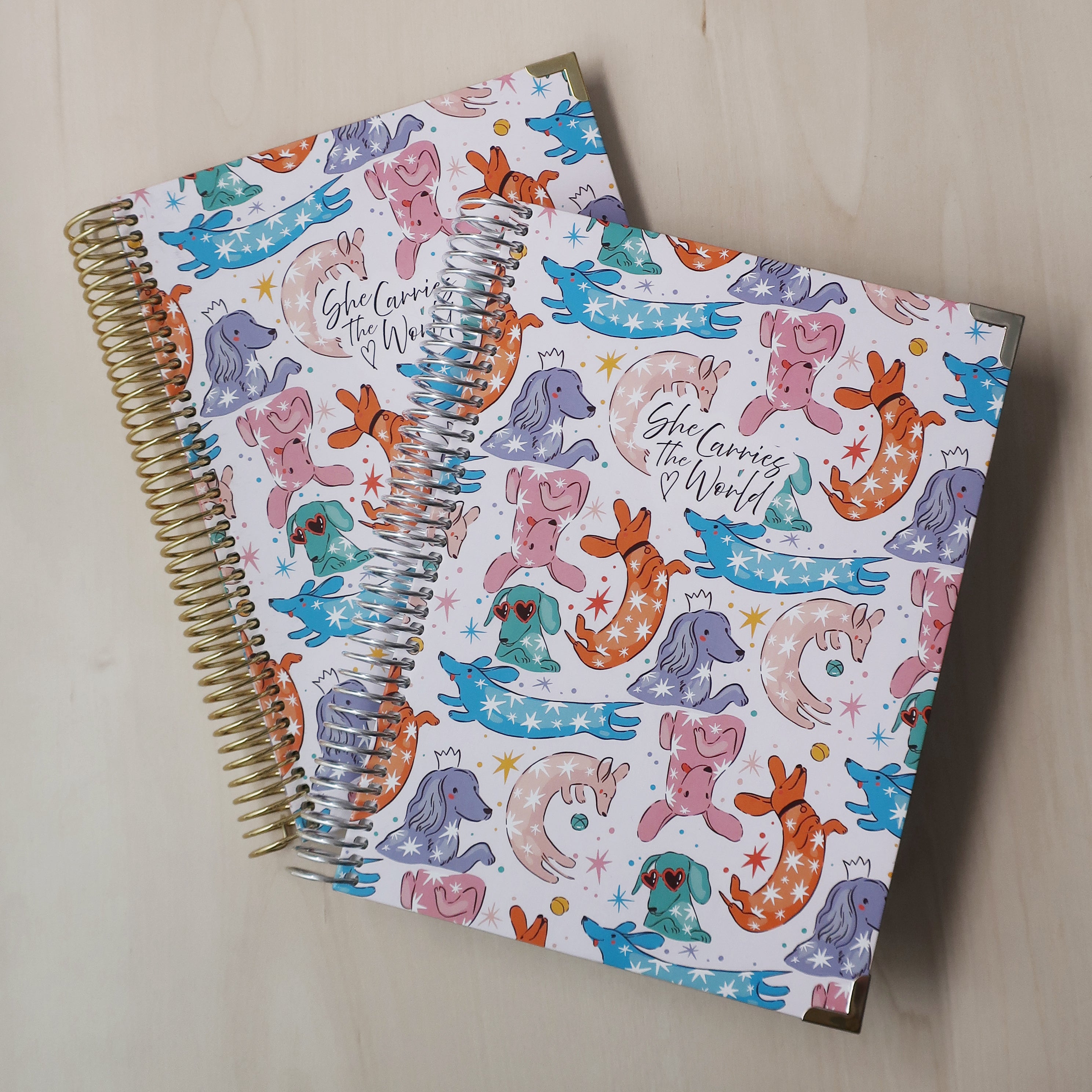Two spiral-bound pregnancy journals with abstract colourful dachshund pattern on a wooden desk. With the 'She Carries The World' logo printed to the cover. 