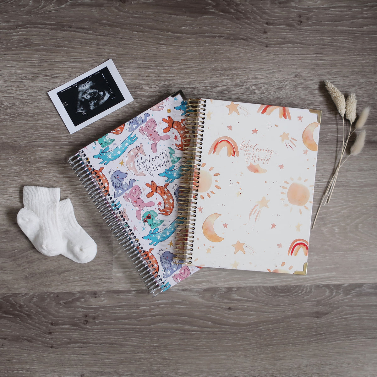 Two Pregnancy Journals On A Wooden Background Surrounded By A Ultrasound & Baby Socks. Ring Bound Pregnancy Journals With Colour Dog Illustration Cover, Soft Watercolour Rainbow Cover.