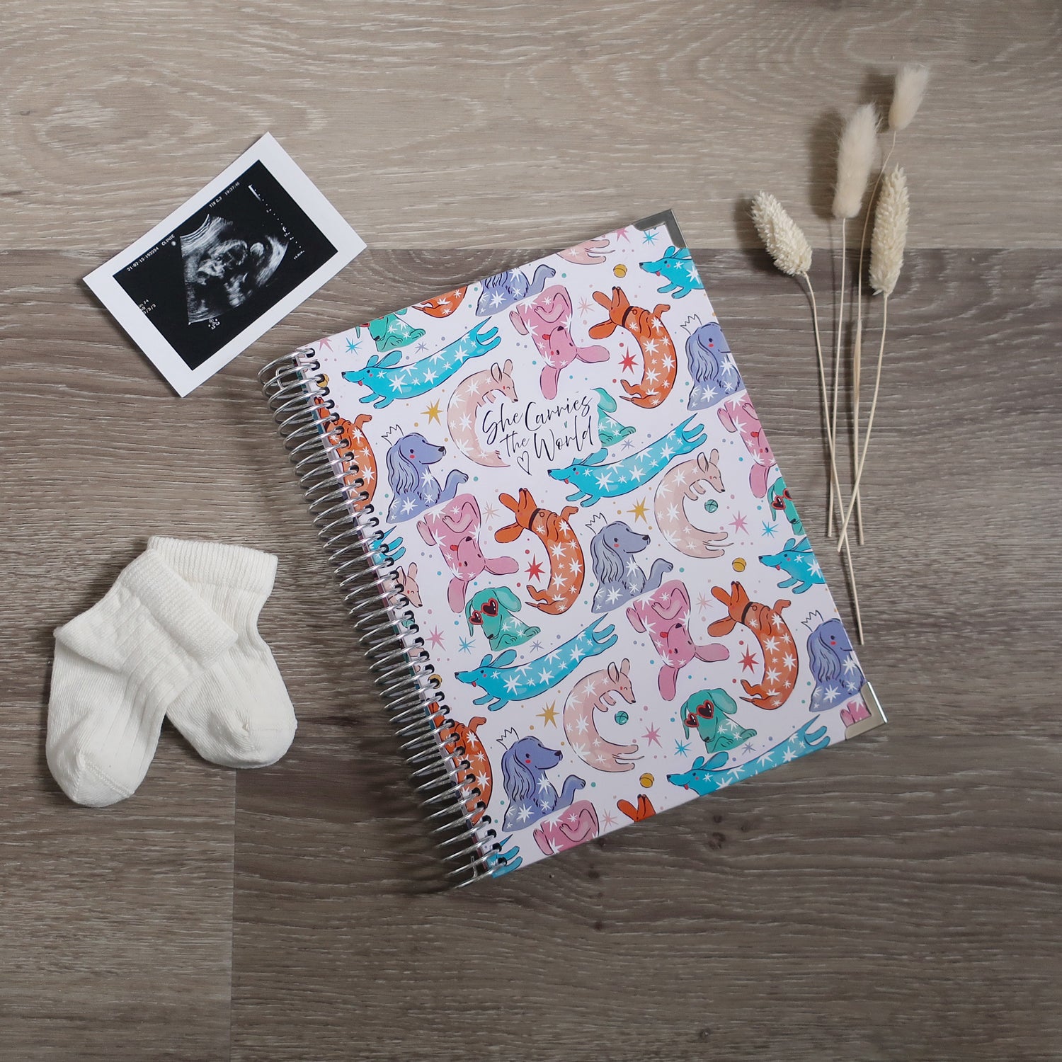 Colourful Dachshund Dog printed spiral-bound pregnancy journal on a wooden desk, next to a ultrasound photo and baby socks.