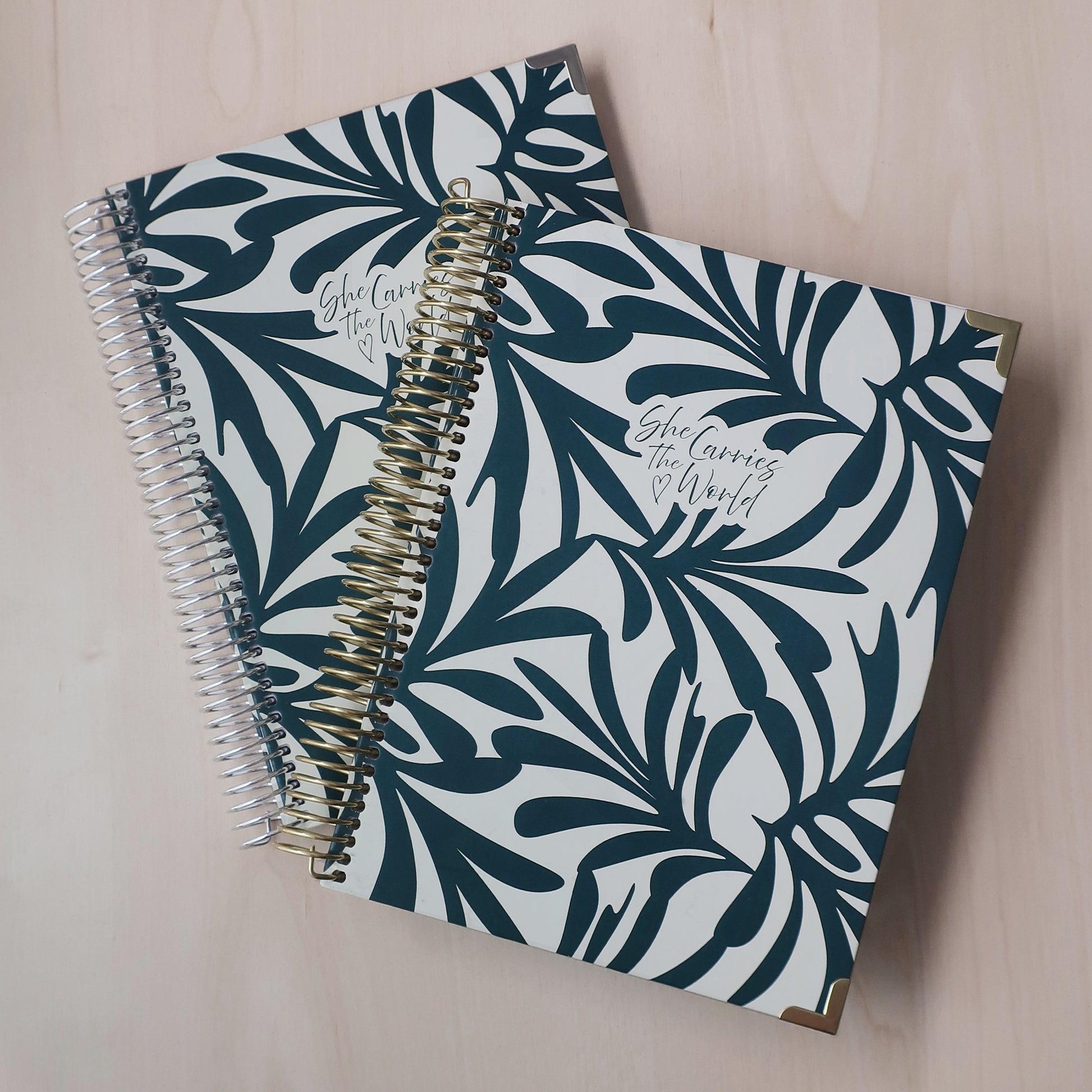Two spiral-bound pregnancy journals with abstract dark green foliage pattern on a wooden desk. With the 'She Carries The World' logo printed to the cover. 