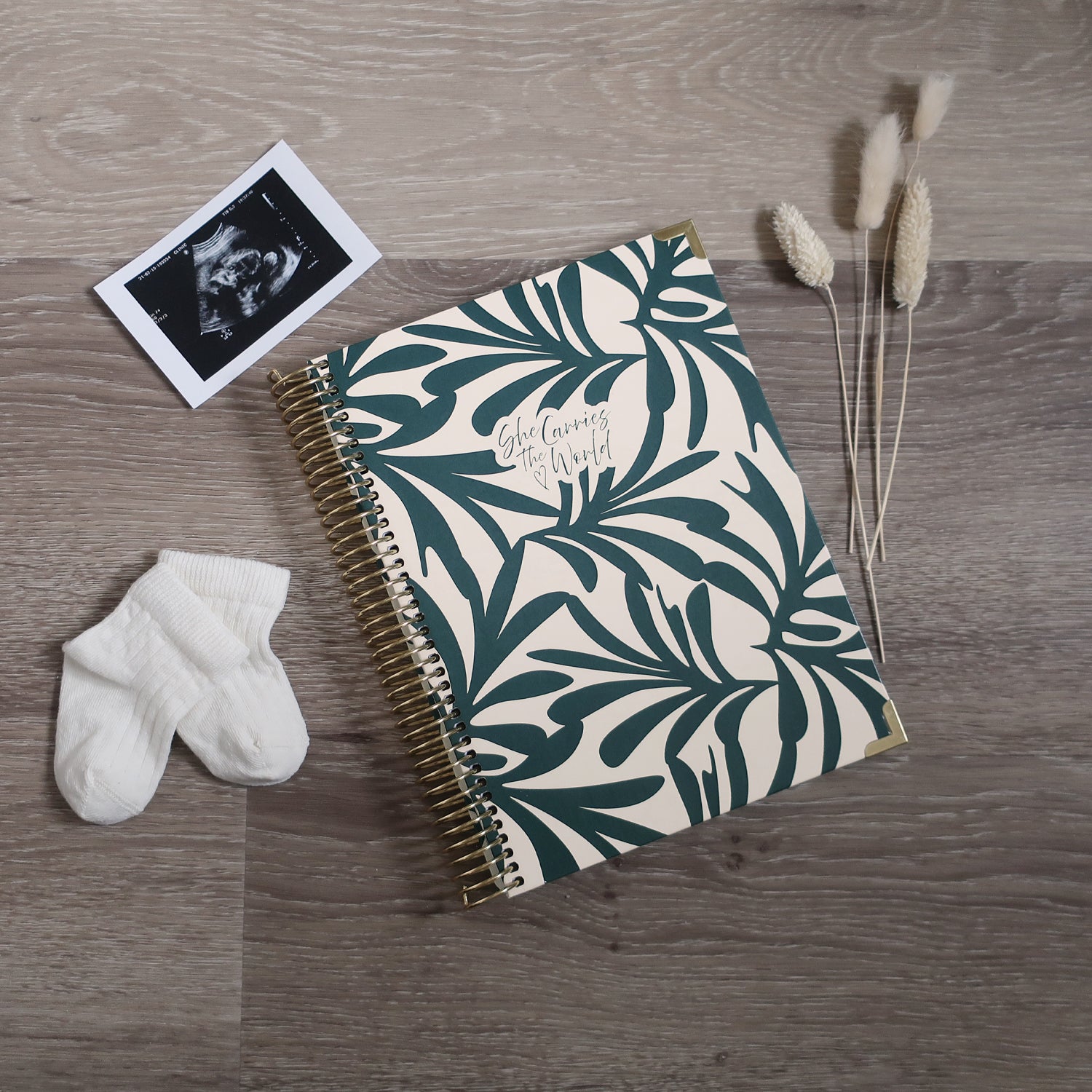Abstract botanical printed spiral-bound pregnancy journal on a wooden desk, next to a ultrasound photo and baby socks.