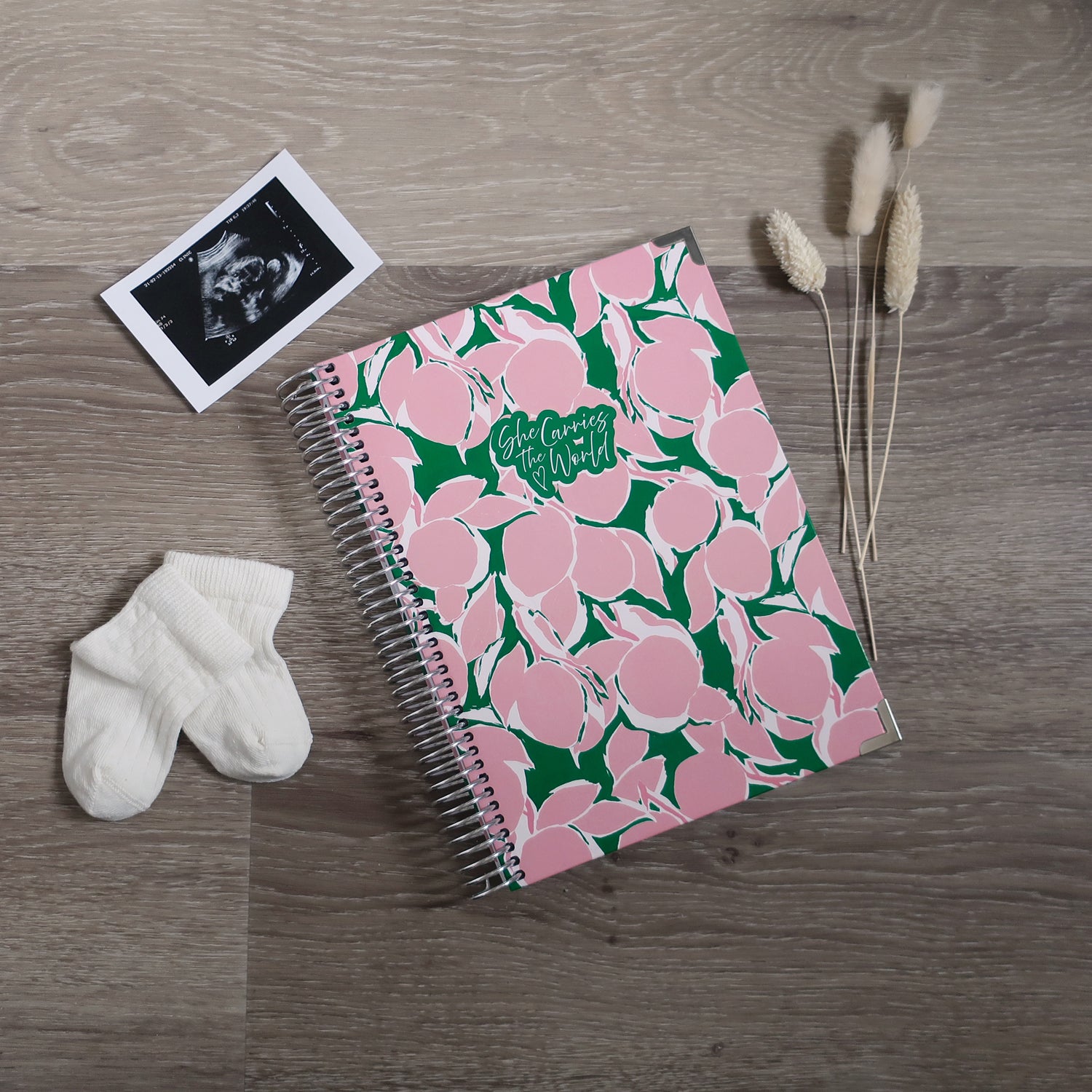 Abstract pink lemons printed spiral-bound pregnancy journal on a wooden desk, next to a ultrasound photo and baby socks.