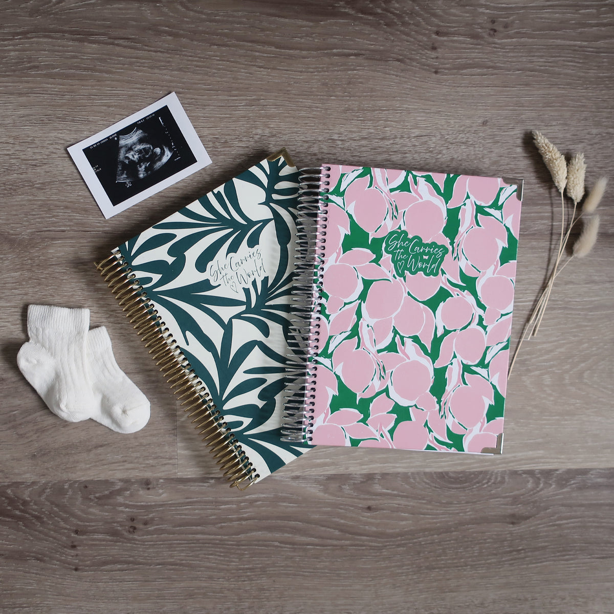 Two Pregnancy Journals On A Wooden Background Surrounded By A Ultrasound & Baby Socks. Ring Bound Pregnancy Journals With Exotic Leaves Cover, Pink Lemons Cover.