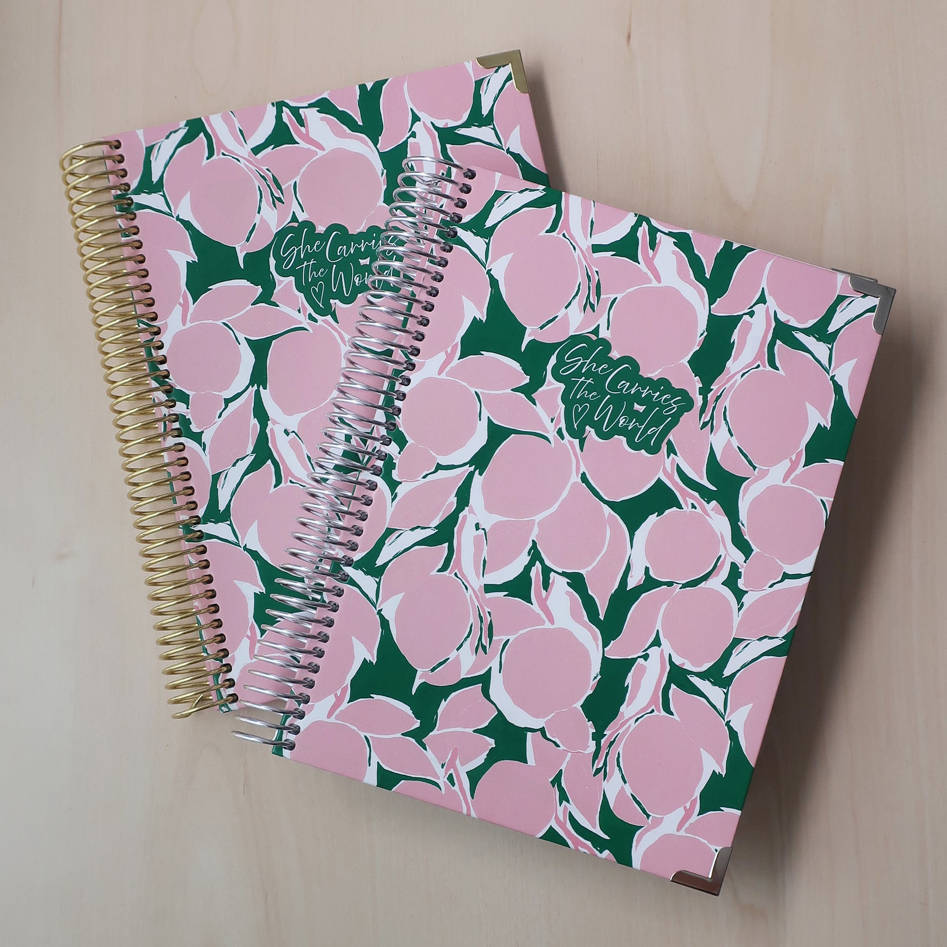 Two spiral-bound pregnancy journals with pink green lemon abstract pattern on a wooden desk. With the 'She Carries The World' logo printed to the cover. 