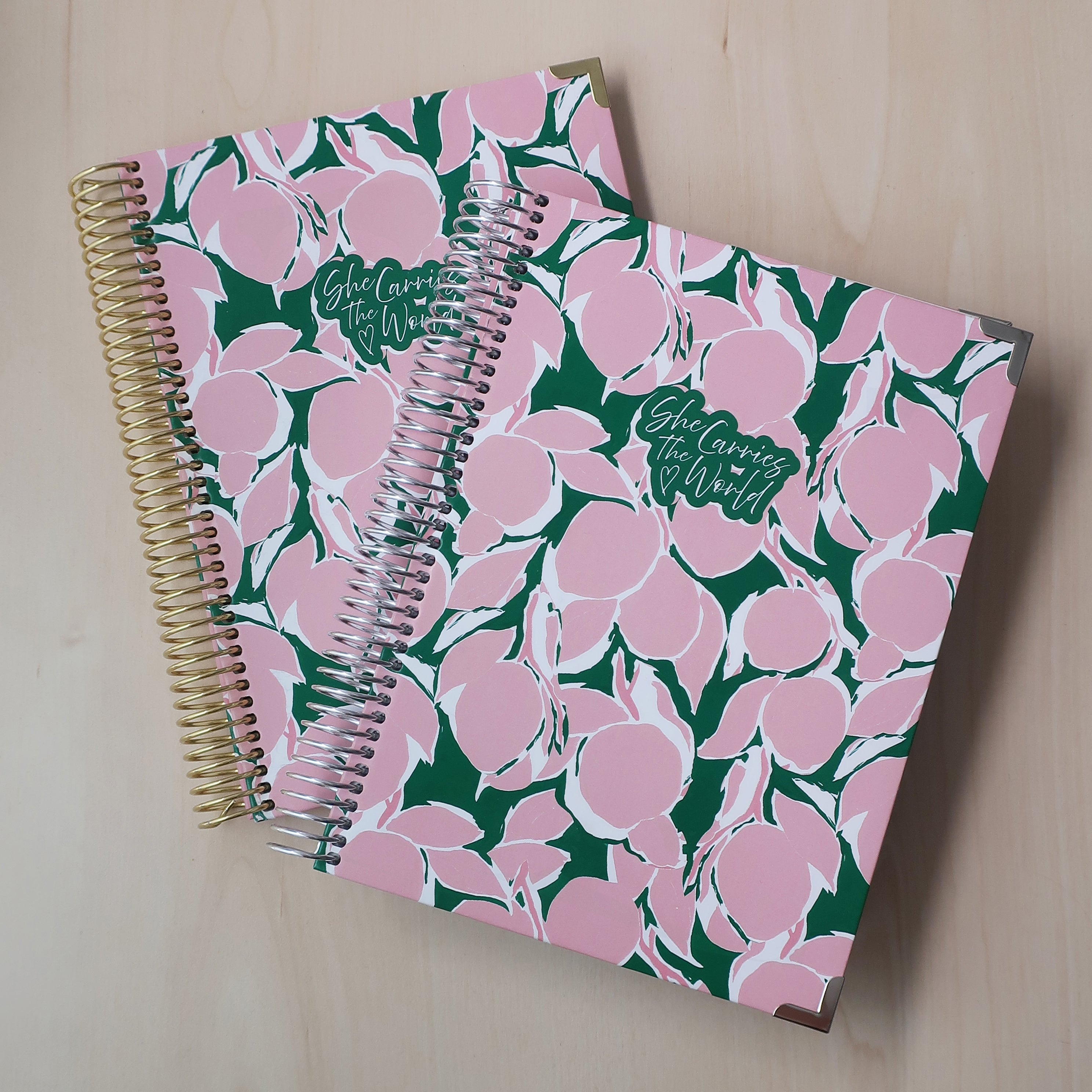 Two spiral-bound pregnancy journals with pink green lemon abstract pattern on a wooden desk. With the 'She Carries The World' logo printed to the cover. 