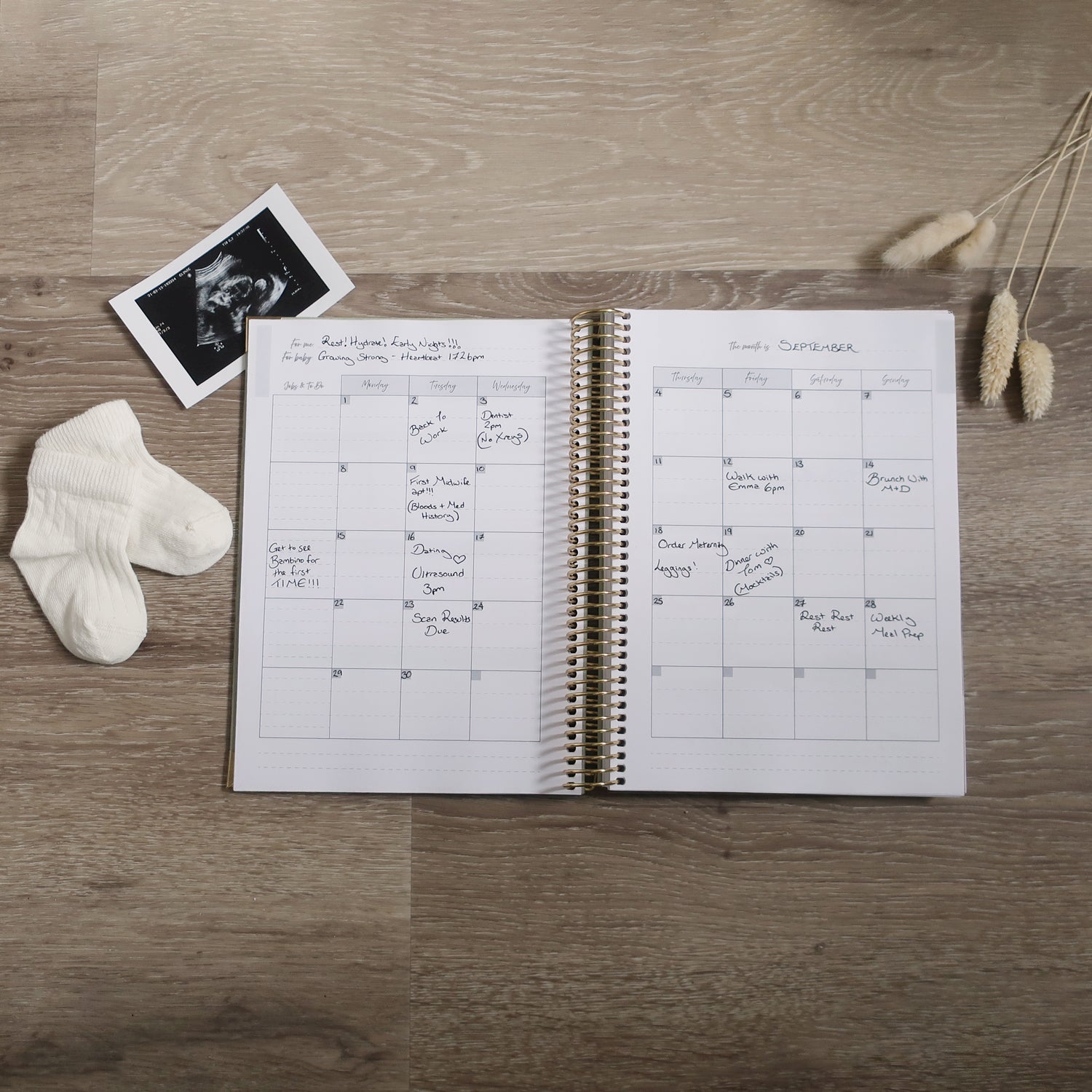 Spiral Bound Pregnancy Journal Open On Monthly Overview Page with ultrasound scan and socks on a wooden desk