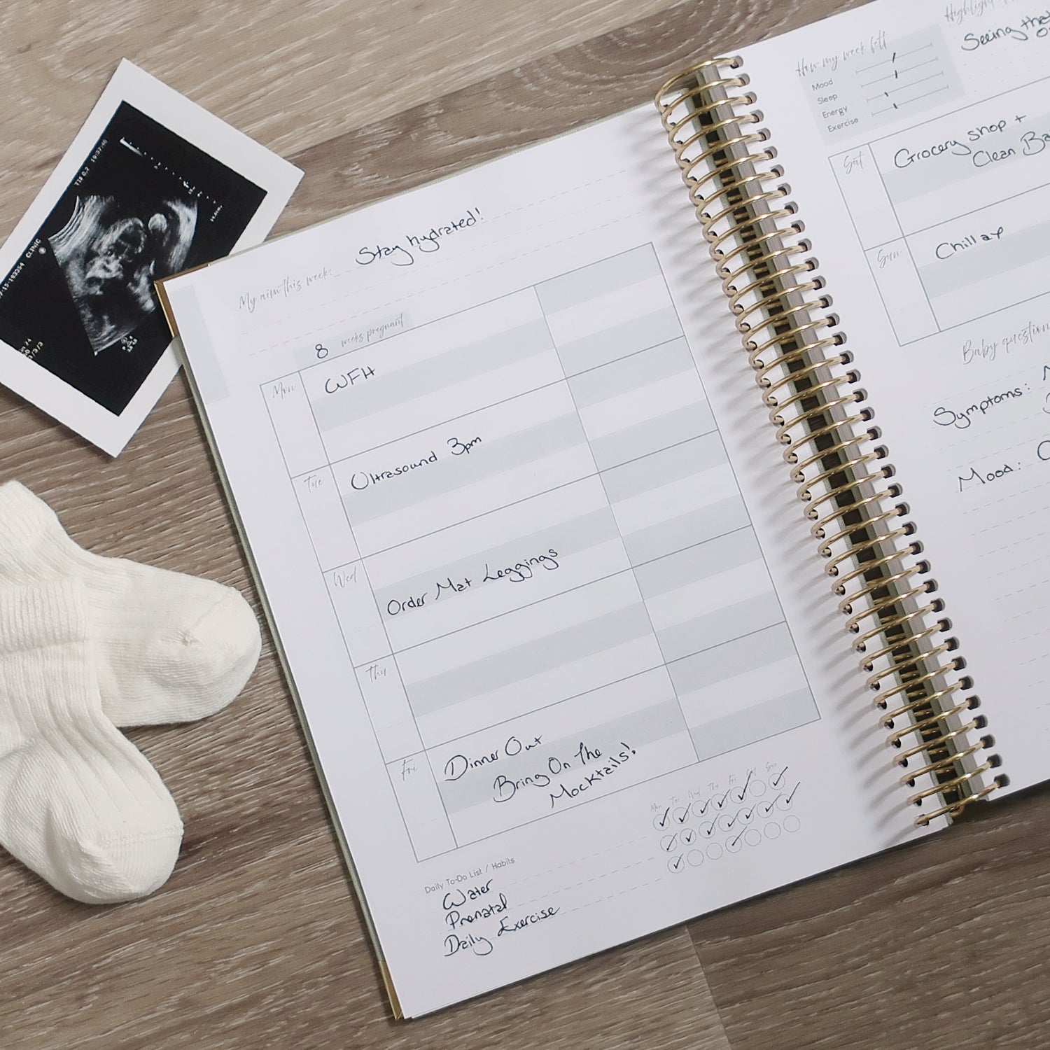 Pregnancy Journal Open On Weekly Overview Page with ultrasound scan and socks on a wooden desk