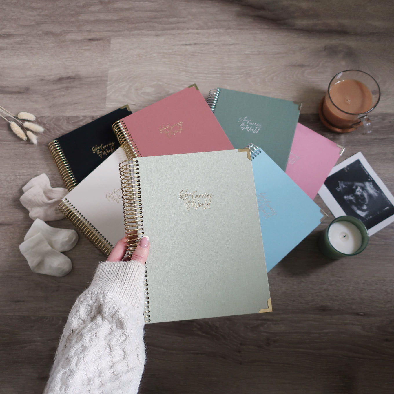 Collection of linen pregnancy journals on a wooden surface with a hand holding one. 'She Carries The World' logo embossed in metallics